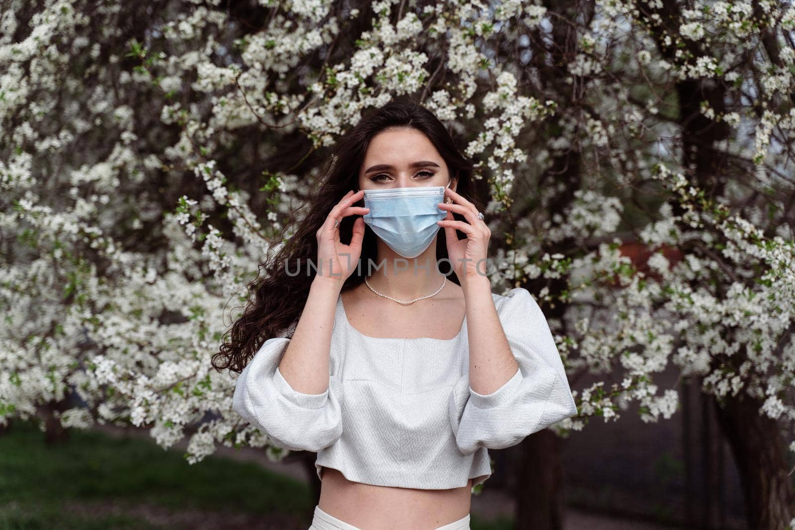 Girl in medical mask near white blooming trees in the park. Outdoor walking countryside at quarantine coronavirus covid-19 period. Spring lifestyle