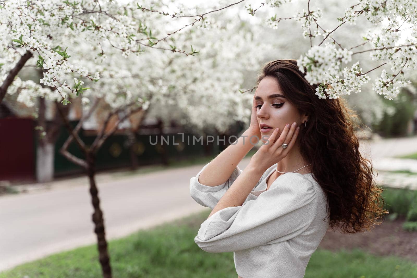 Spring lifestyle. Model posing near white blooming trees without mask outdoor countryside. Dreaming girl with curly hair