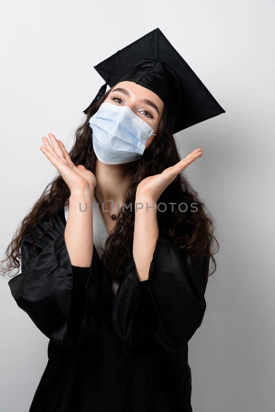 Surprised bachelor girl in medical mask at coronavirus covid-19 period. Master in graduation robe and cap on white background. Distance learning online. Study at home. Graduation from college by Rabizo