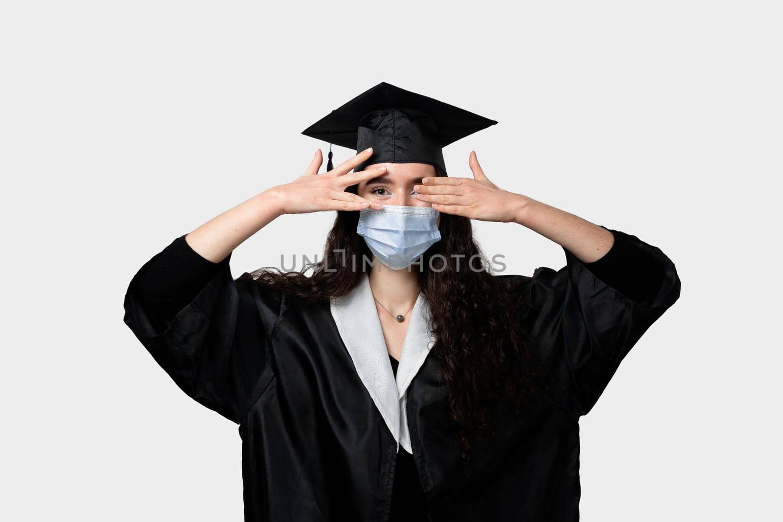 Surprised bachelor girl in medical mask at coronavirus covid-19 period. Master in graduation robe and cap on white background. Distance learning online. Study at home. Graduation from college.