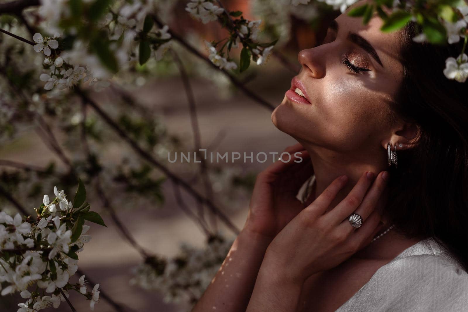 The sun shines on the girl's face near the flowering trees in the park. Portrait of attractive girl with curly hair in the garden