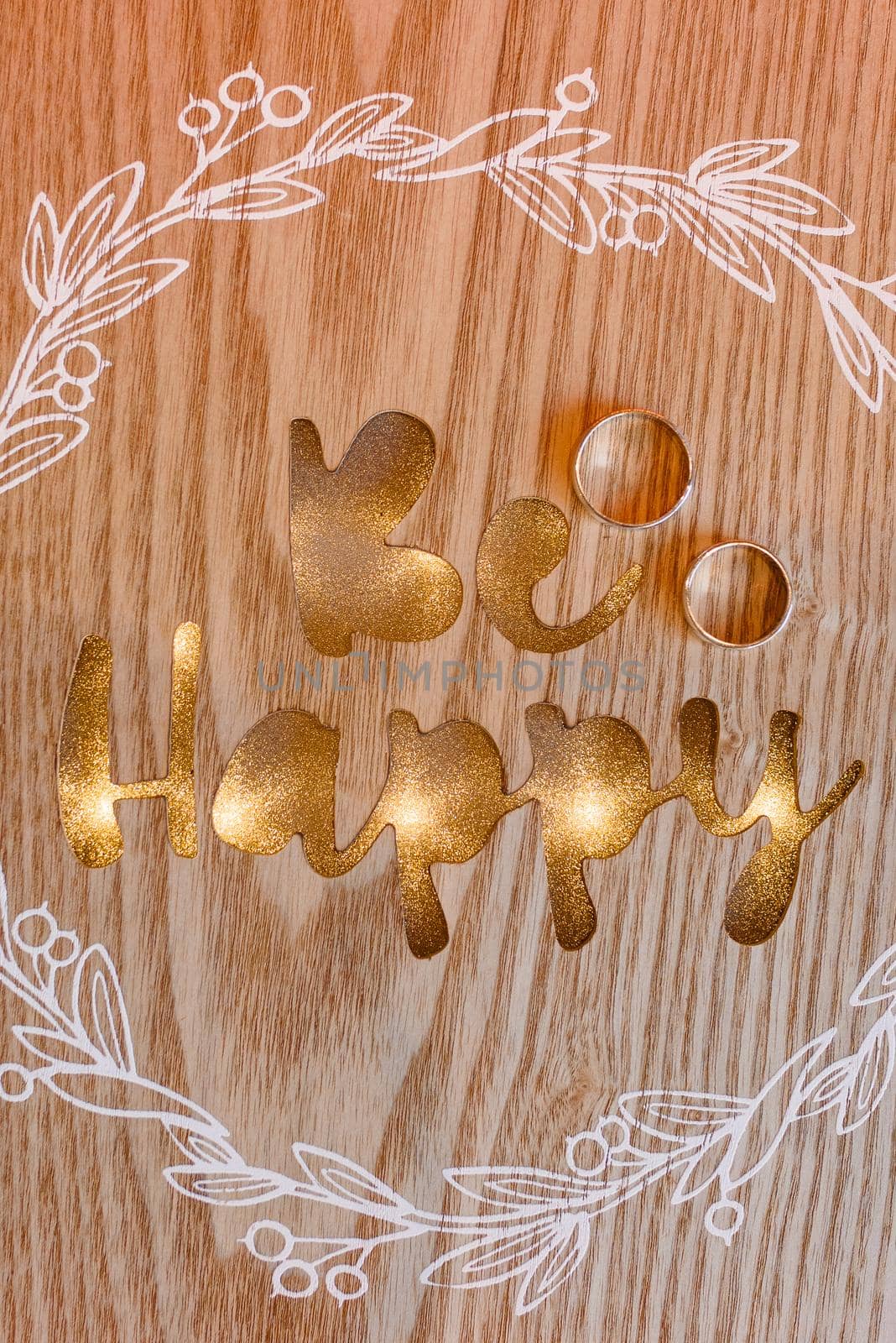 Wedding rings on a wooden background with golden inscription be happy and white pattern.