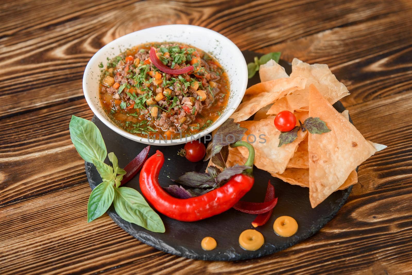 Mexican dish with meat, chili, tomato, corn with nachos in a white plate on a black slate platter, garnished with basil, blue onions and cherry tomatoes. Spicy Mexican dish on a wooden table. by Rabizo