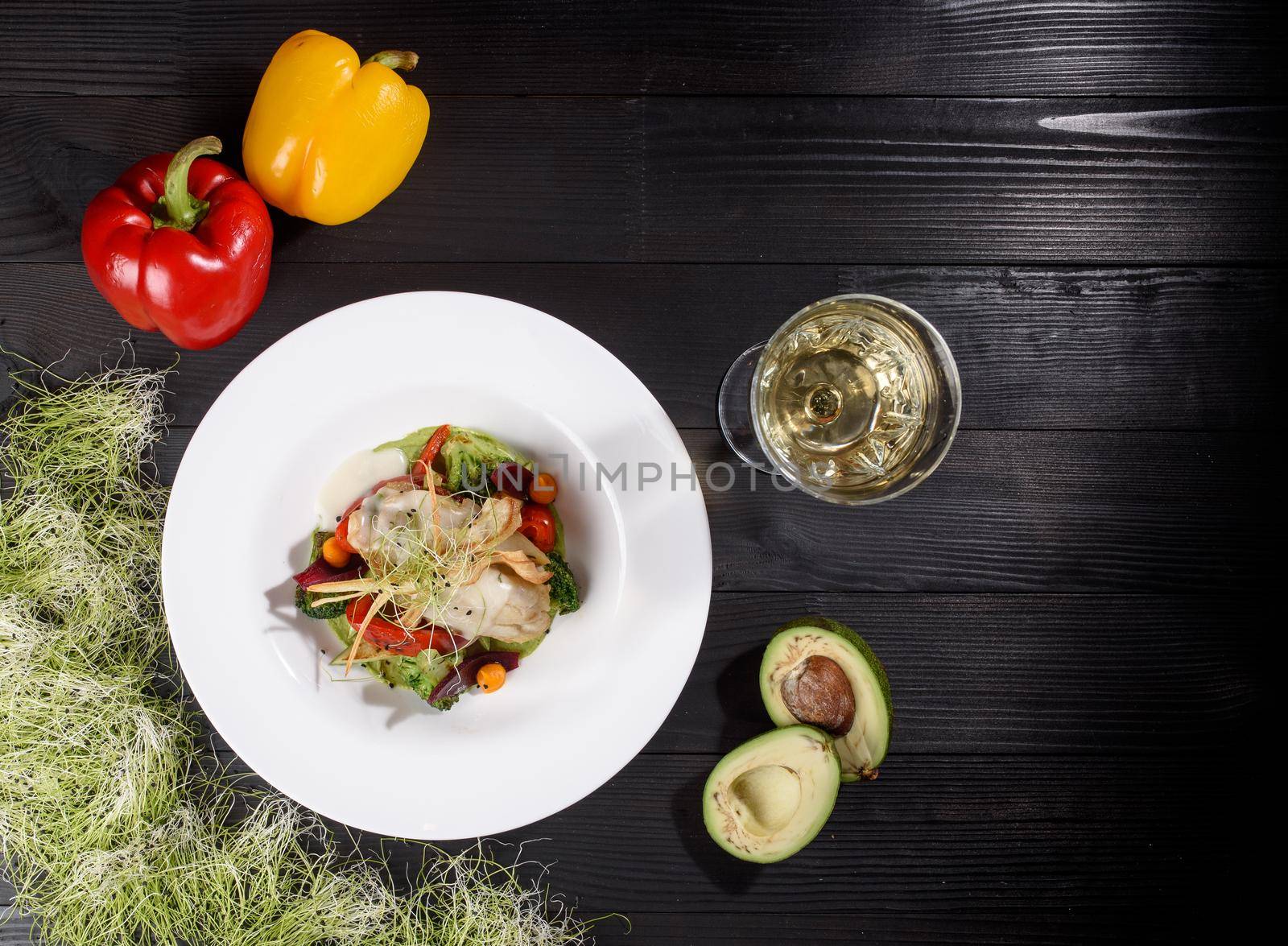 Chicken fillet with white sauce with grilled vegetables, garnished with microgreens on a white plate against the background of a glass of white wine, bell pepper and avocado on a wooden table.
