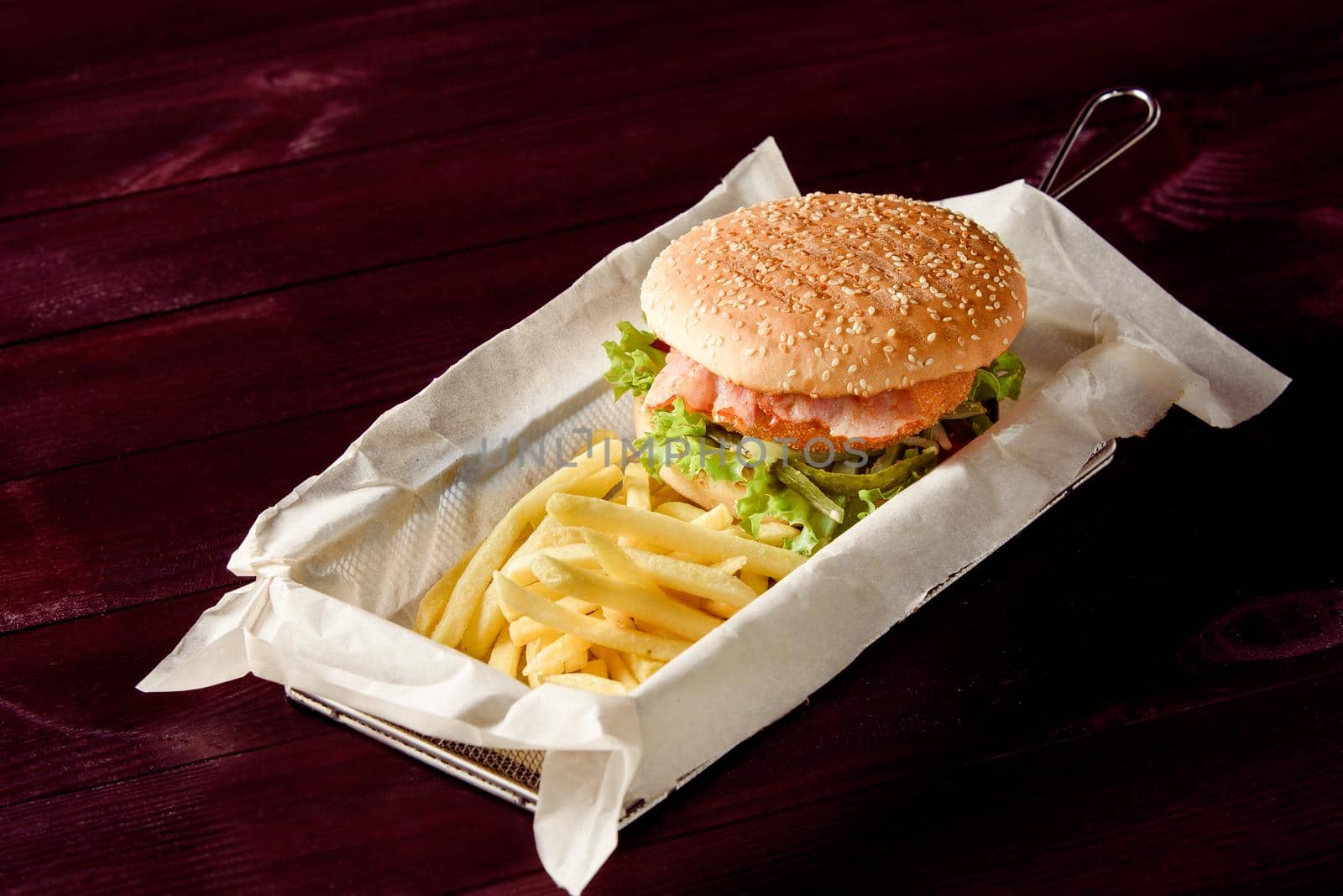 Burger with cutlet, bacon, salad, pickled cucumber and potato fries on parchment in a metal basket on a wooden background.