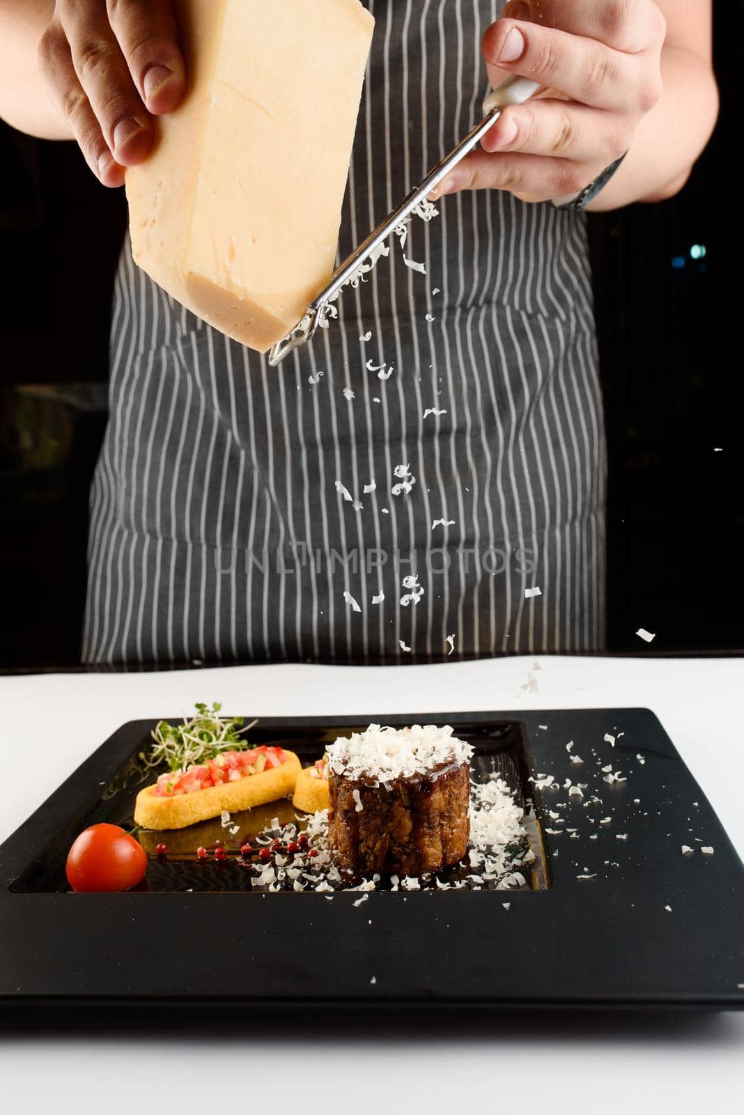 Сhef grates cheese on a gourmet restaurant dish with meat and vegetables on a black square plate.