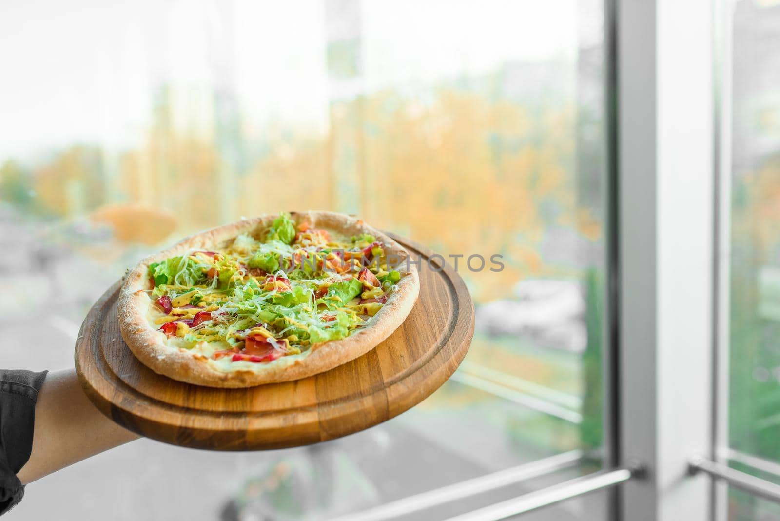Delicious fresh italian pizza with ham, salami, tomatoes, salad and parmesan on a wooden board against the background of a window.
