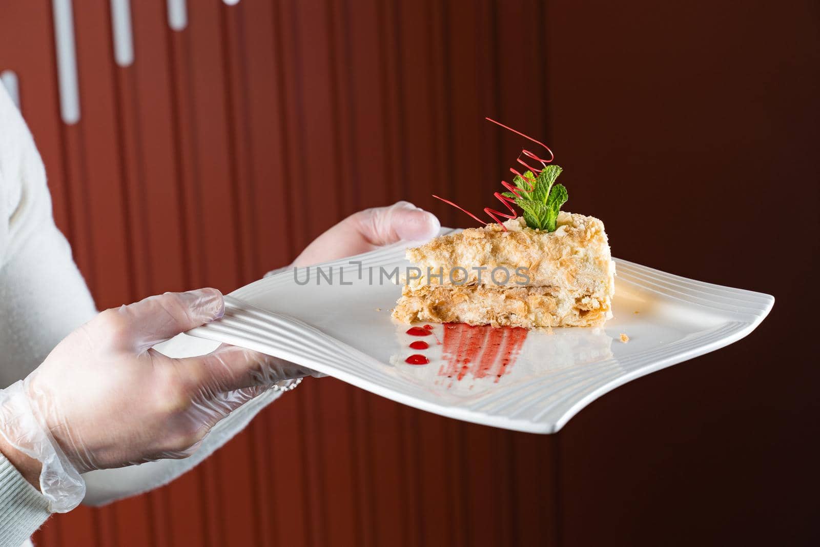 A waiter in gloves holds a white plate of napoleon cake decorated with a spiral of red chocolate, mint and berry jam on a red background.