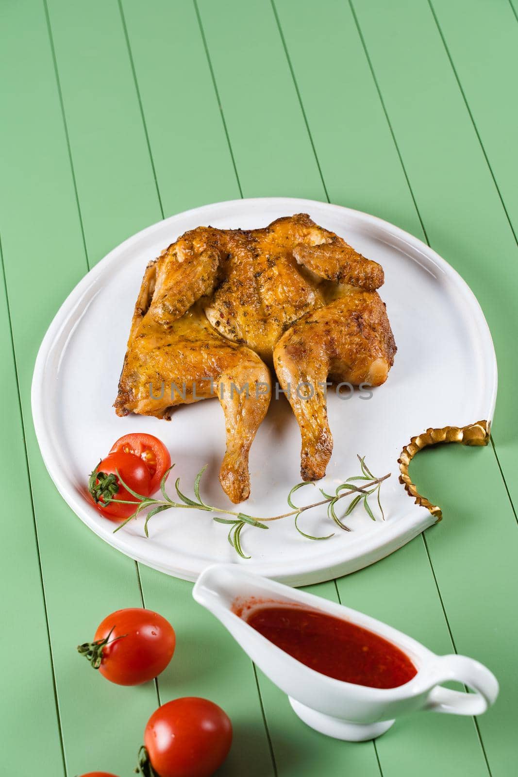 Chicken tobacco with tomato sauce, rosemary and tomatoes on a beautiful white plate on a green background. Grilled chicken.