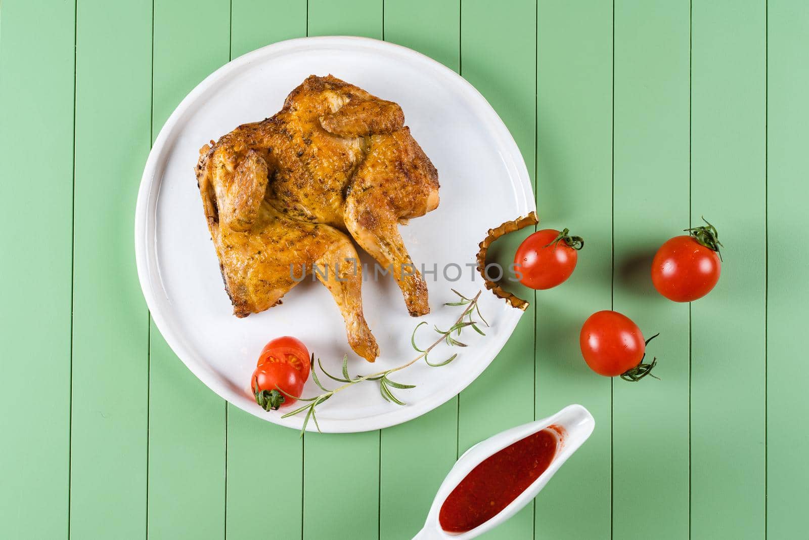 Chicken tobacco with tomato sauce, rosemary and tomatoes on a beautiful white plate on a green background. Grilled chicken.