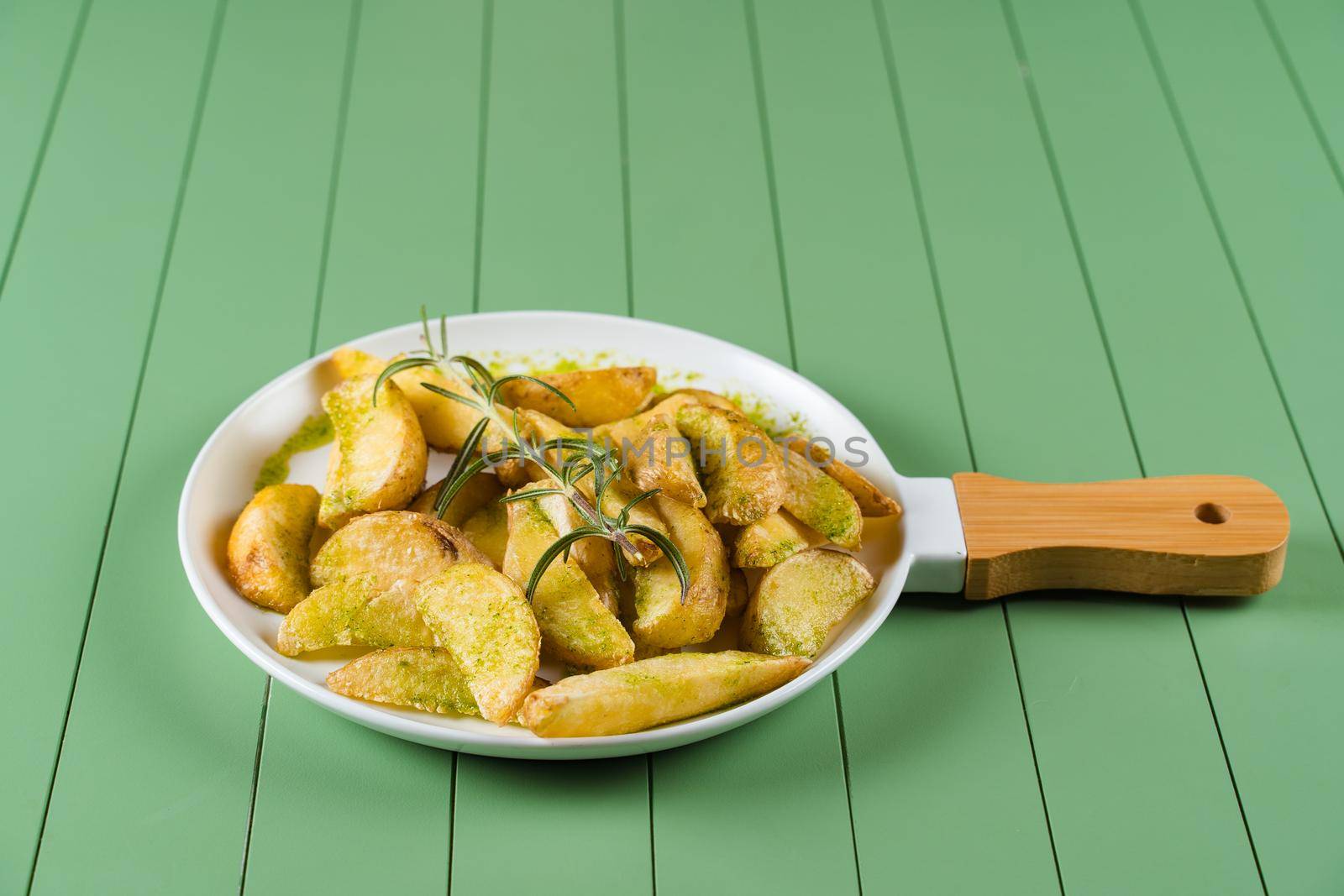 Baked potatoes with green sauce and rosemary on a white plate in the form of a frying pan. Fried potatoes with pesto sauce on a green table by Rabizo