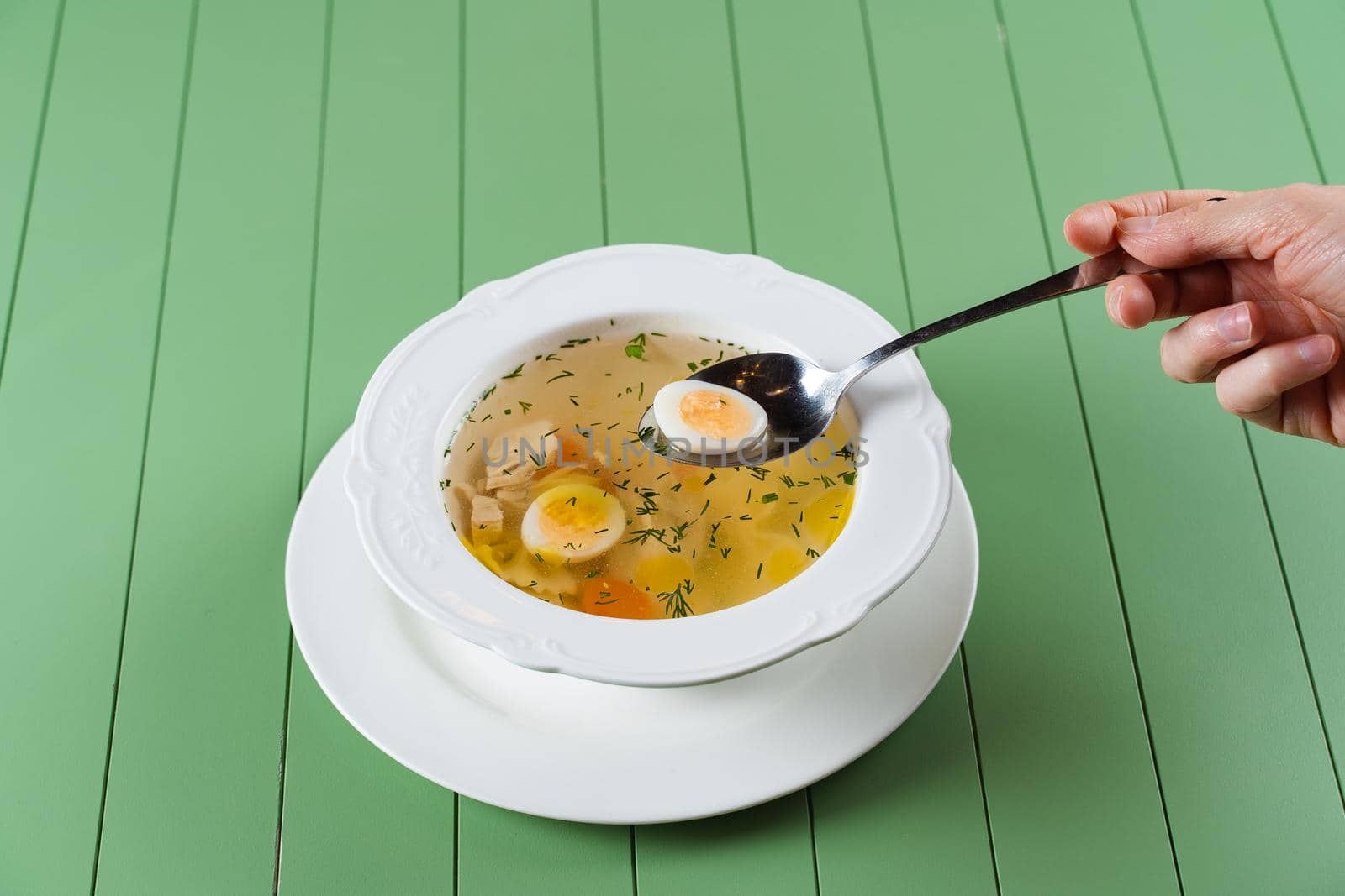 Chicken broth with meat, carrots, herbs and quail egg in a white plate on a green background