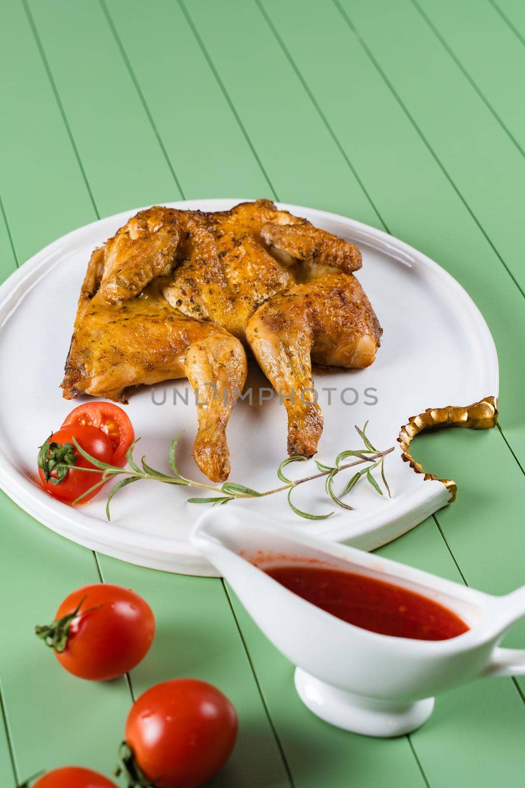 Chicken tobacco with tomato sauce, rosemary and tomatoes on a beautiful white plate on a green background. Grilled chicken by Rabizo