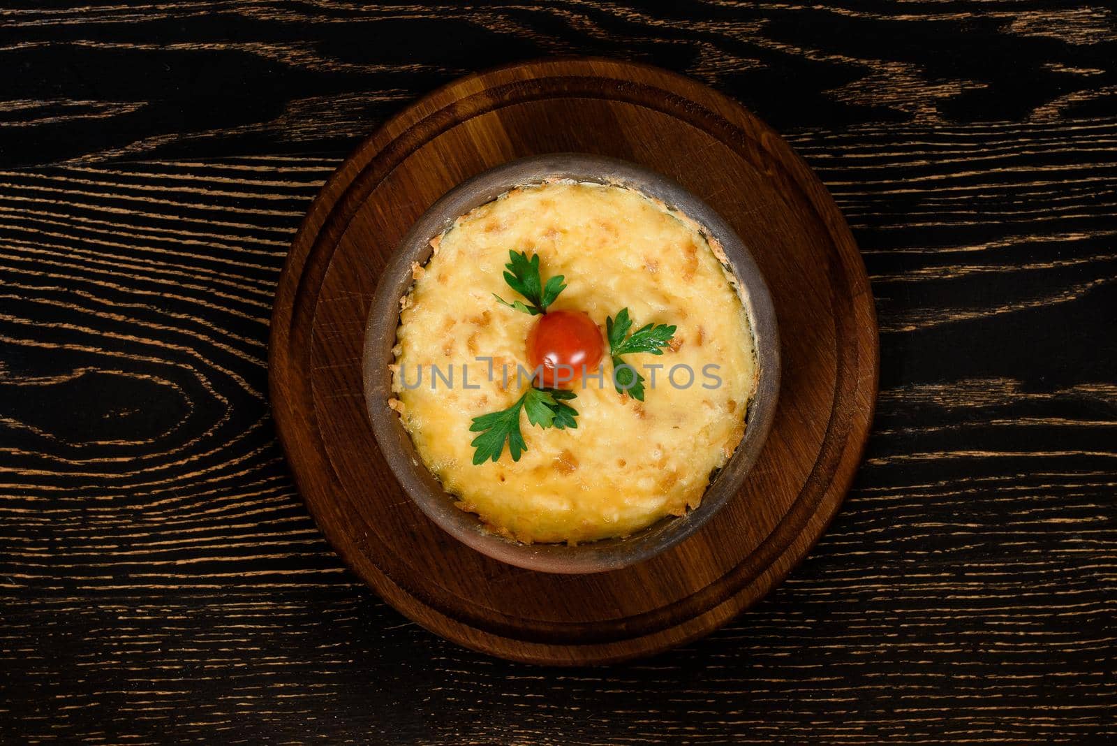 Dish baked with cheese, decorated with cherry tomato and parsley in a clay plate on a wooden board on a dark wooden table by Rabizo