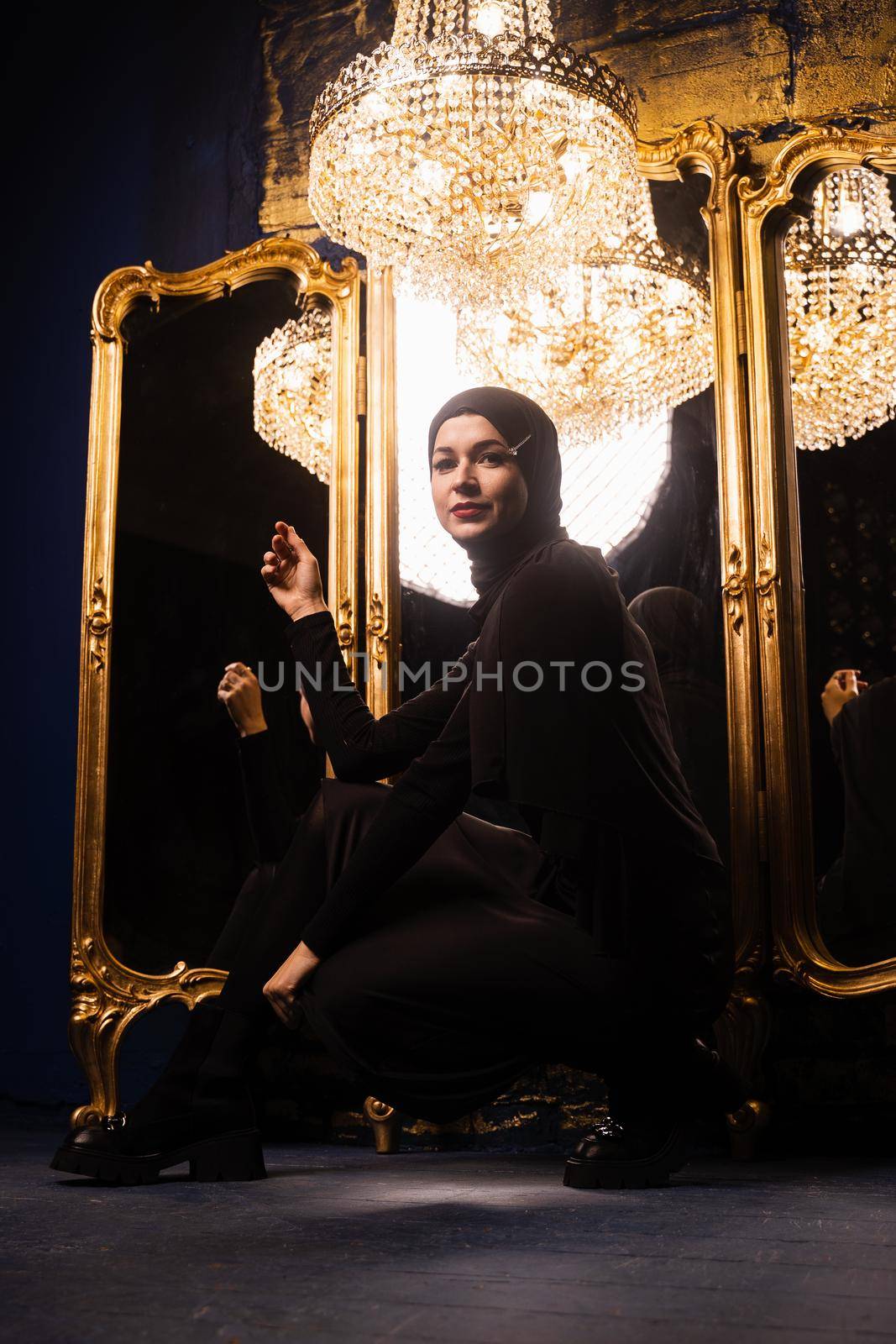 Muslim girl in black hijab near mirrors with her reflections. Fashion muslim model near big expensive chandelier. Islamic religion