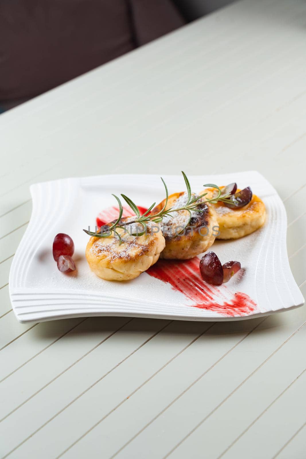 Cheesecakes decorated with powdered sugar, rosemary branch and grapes with berry jam on a white plate. Delicious cottage cheese breakfast.