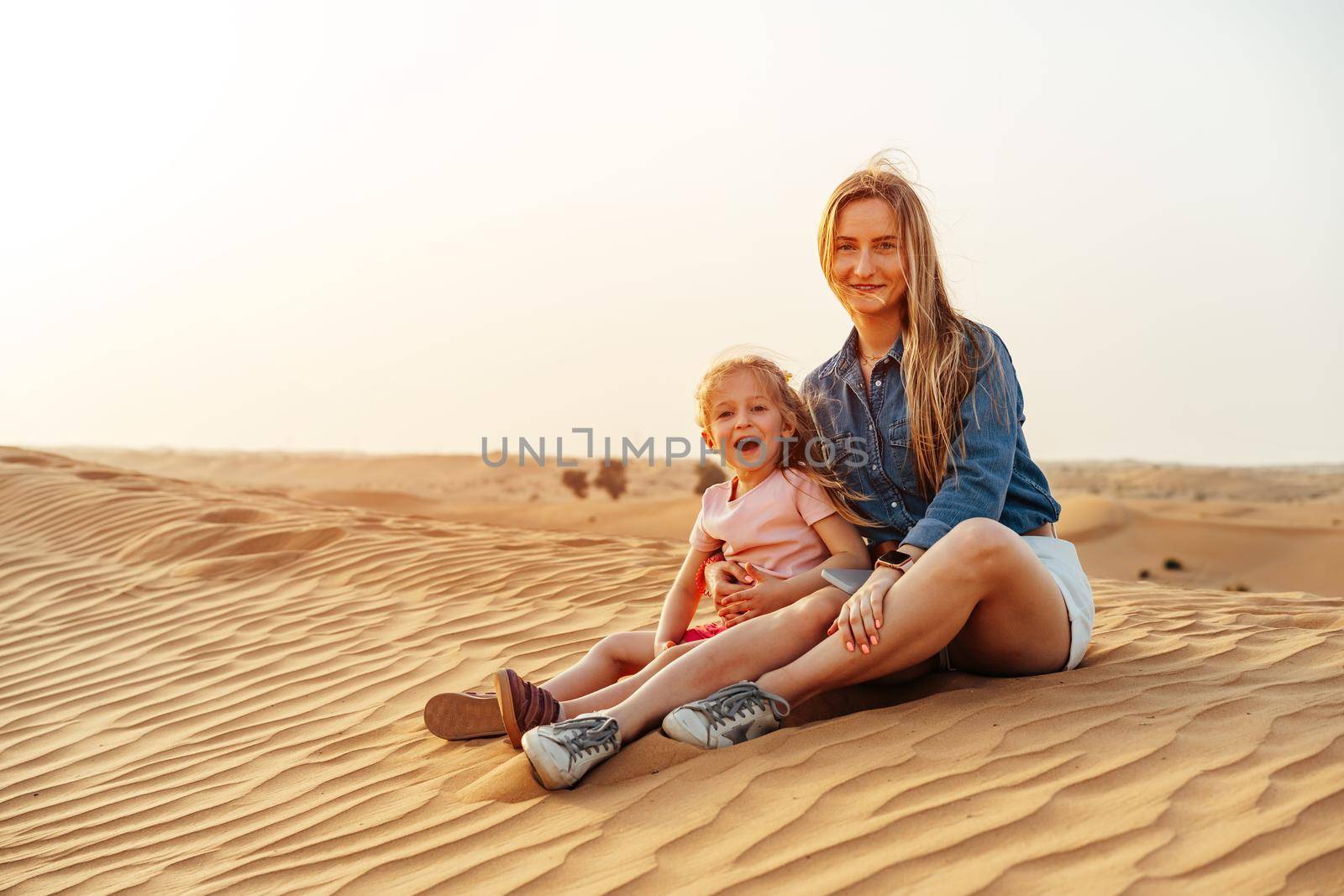 Mother and daughter sitting together on sand dune in the Dubai desert, travel with children concept