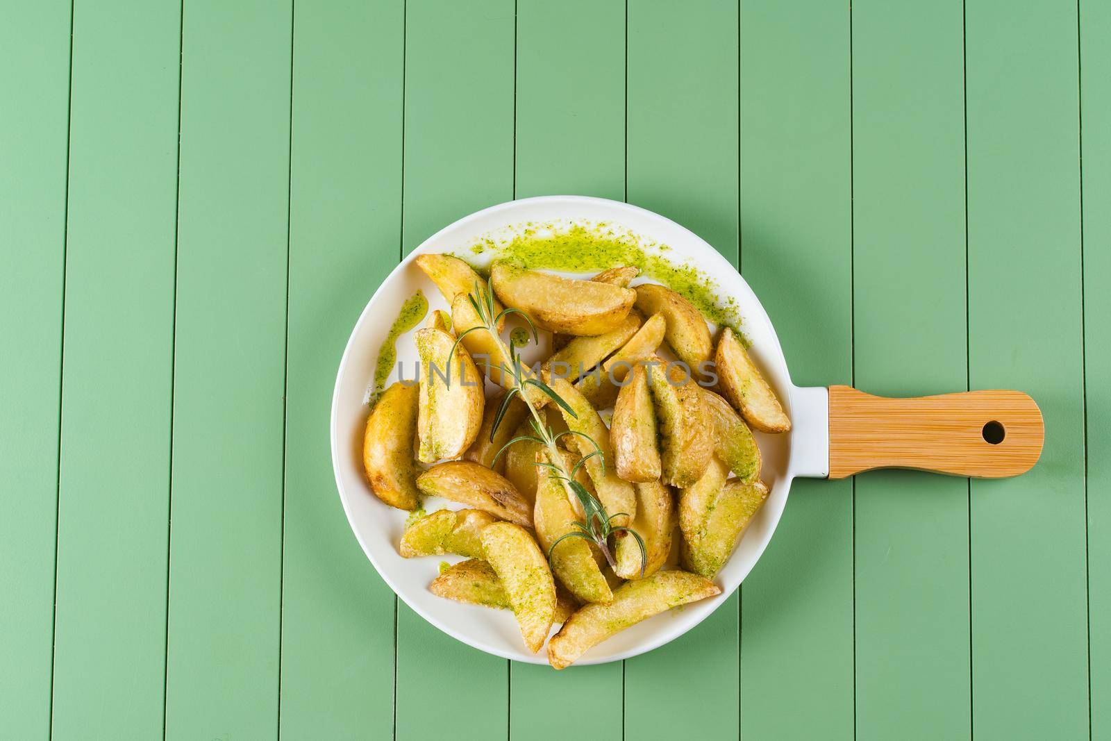 Baked potatoes with green sauce and rosemary on a white plate in the form of a frying pan. Fried potatoes with pesto sauce on a green table by Rabizo