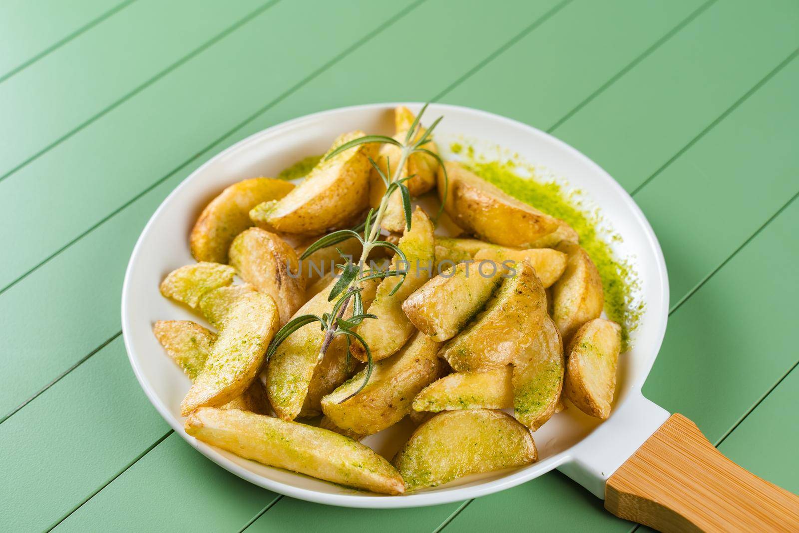 Baked potatoes with green sauce and rosemary on a white plate in the form of a frying pan. Fried potatoes with pesto sauce on a green table.