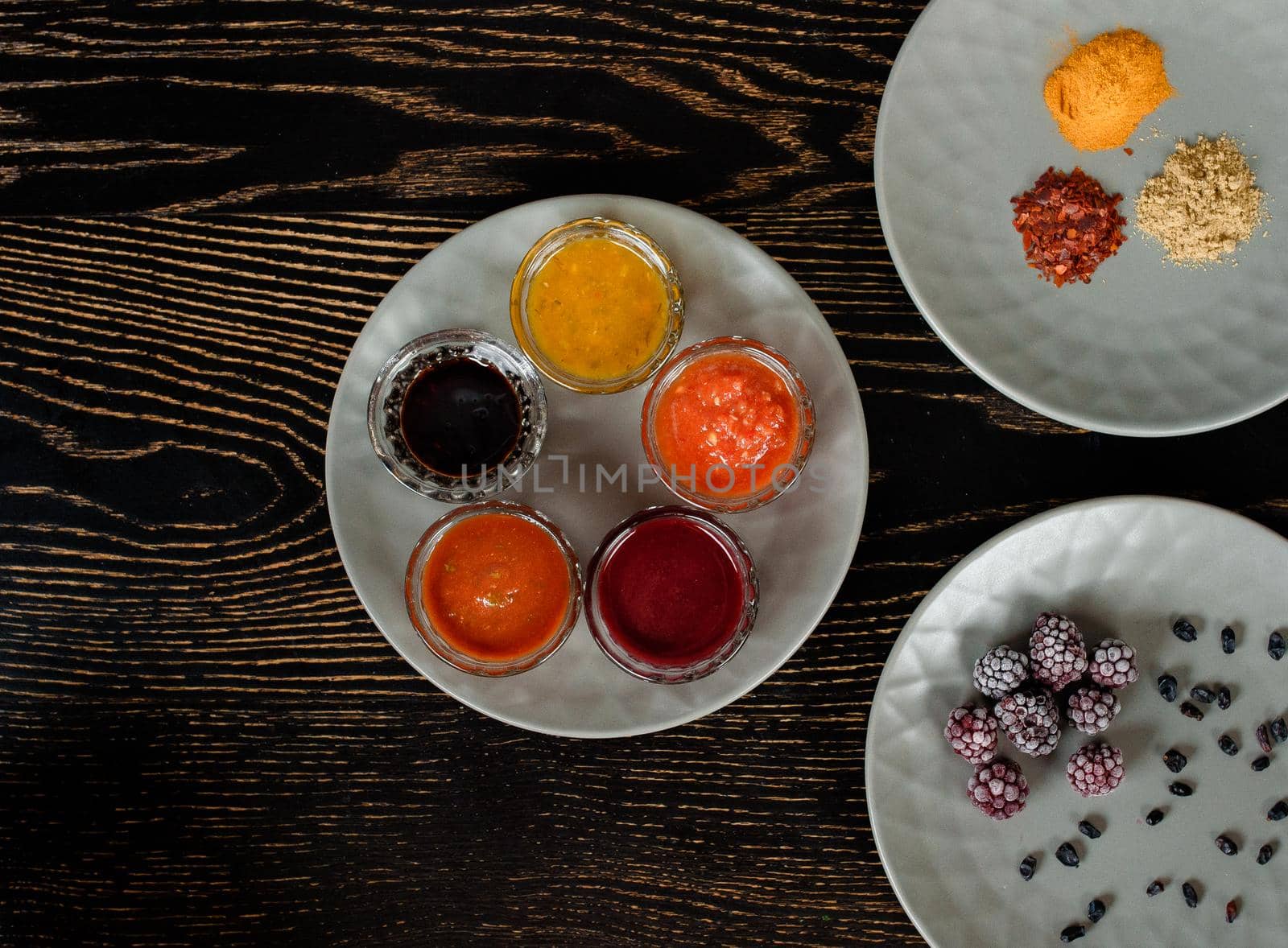Assortment of sauces, spices, frozen berries on gray plates on a dark wooden table. View from above.