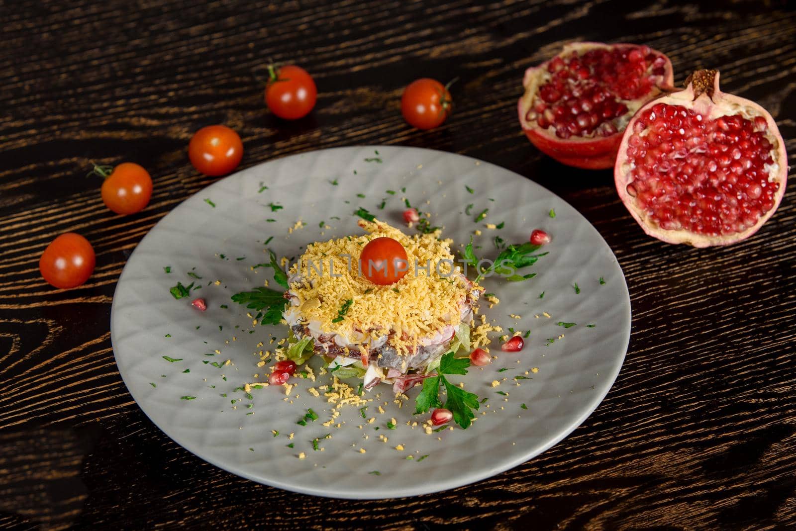 Puff salad with meat, vegetables, cheese, eggs, garnished with herbs and pomegranate and cherry tomatoes on a gray plate. Against the background of halves of pomegranate and cherry tomatoes. by Rabizo