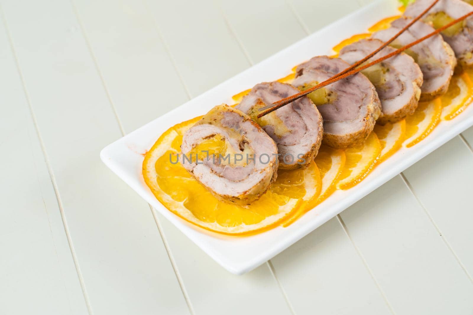 Meat rolls on orange slices on a rectangular white plate on a white background. Sliced pork appetizer.