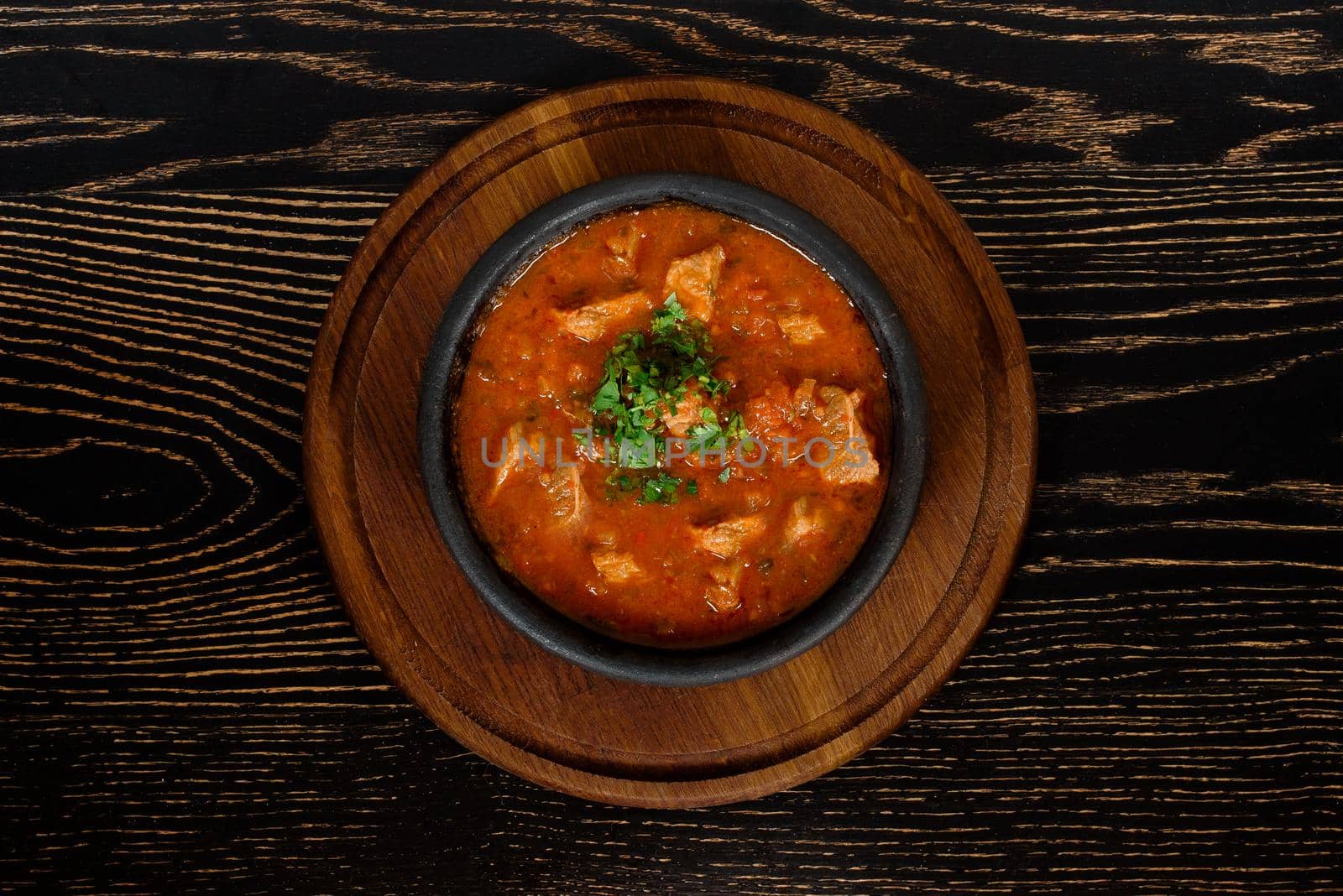 Meat in tomato sauce in a black clay plate on a wooden board on a dark wooden table.