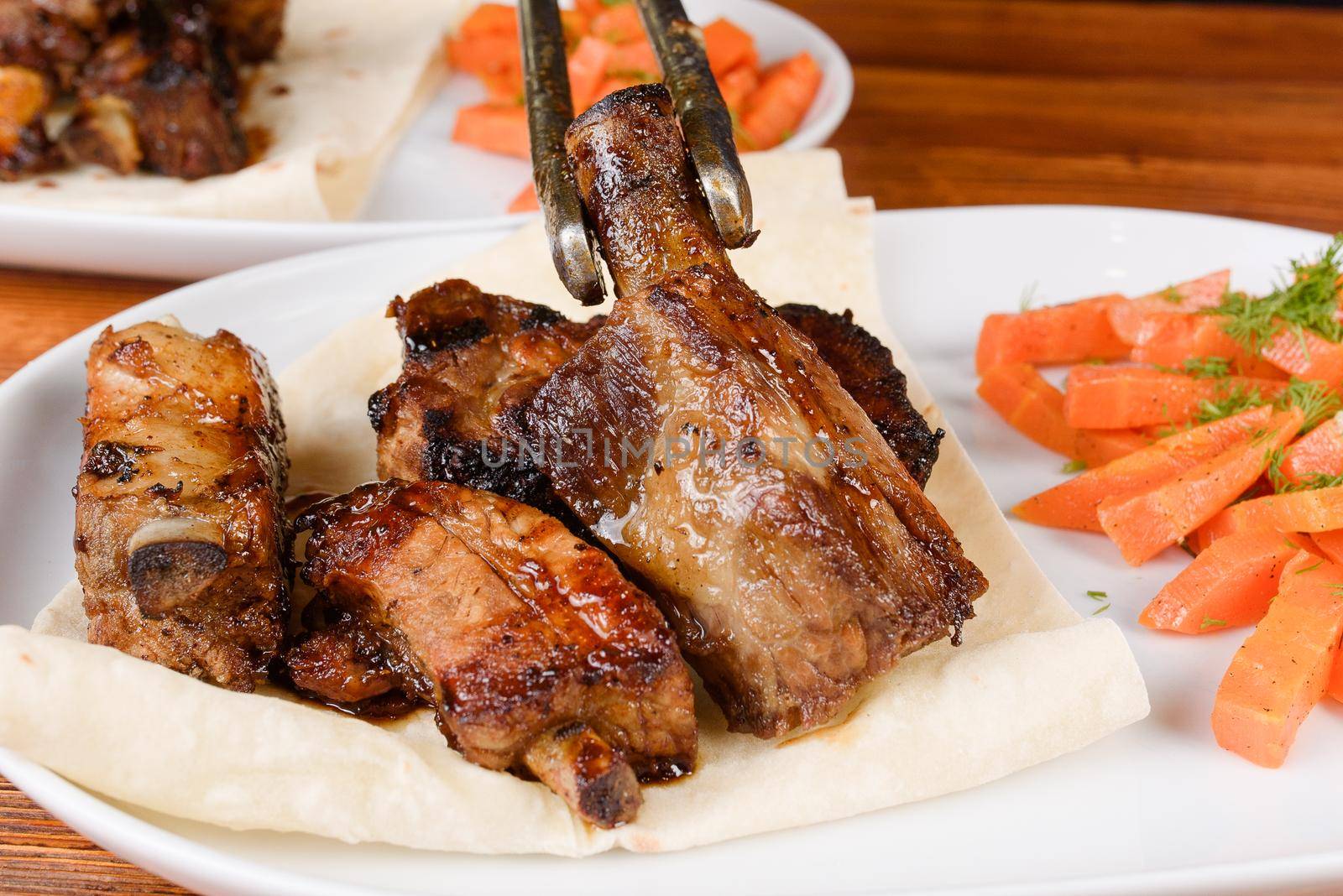 Sliced grilled pork ribs on pita bread with grilled carrots on a white plate on a wooden background.