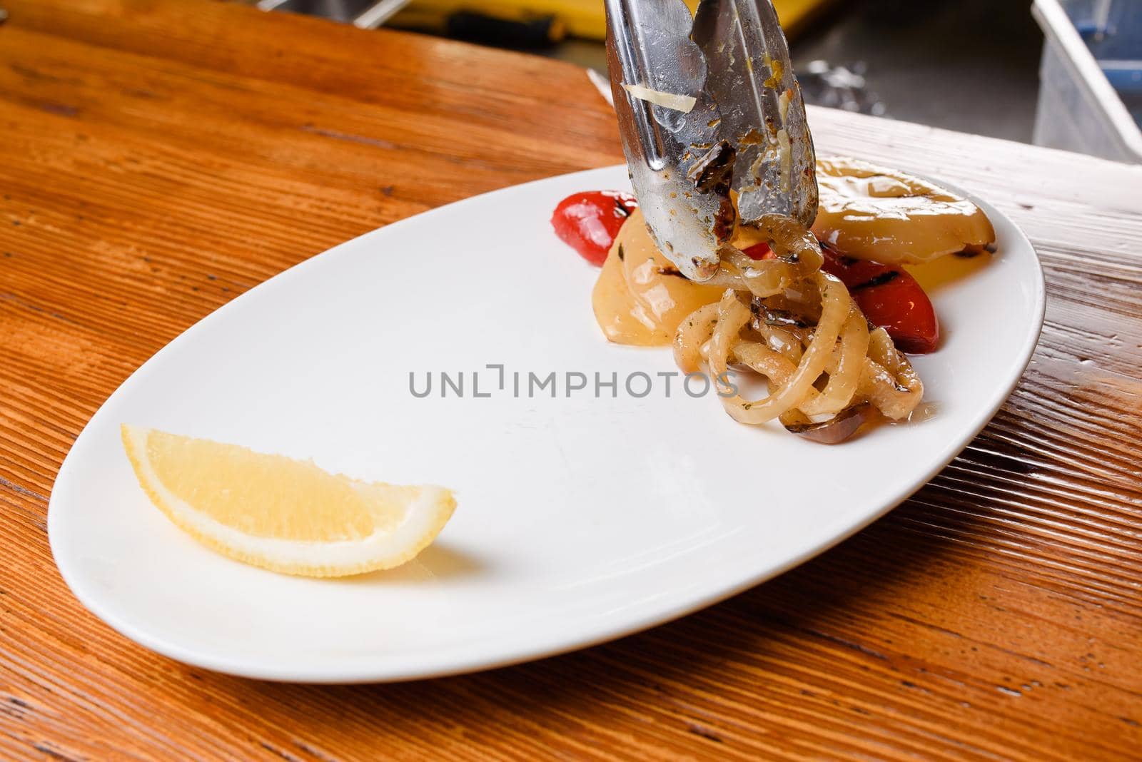 Grilled pepper and onion with lemon on a white plate on a wooden table. Serving the dish with metal tongs.