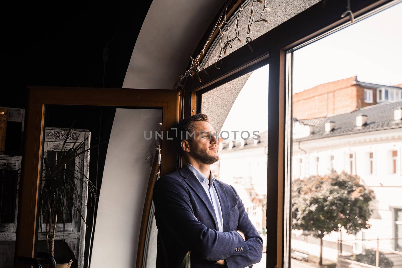 Handsome male model is posing in cafe. Bearded man weared suit stand and smile near window