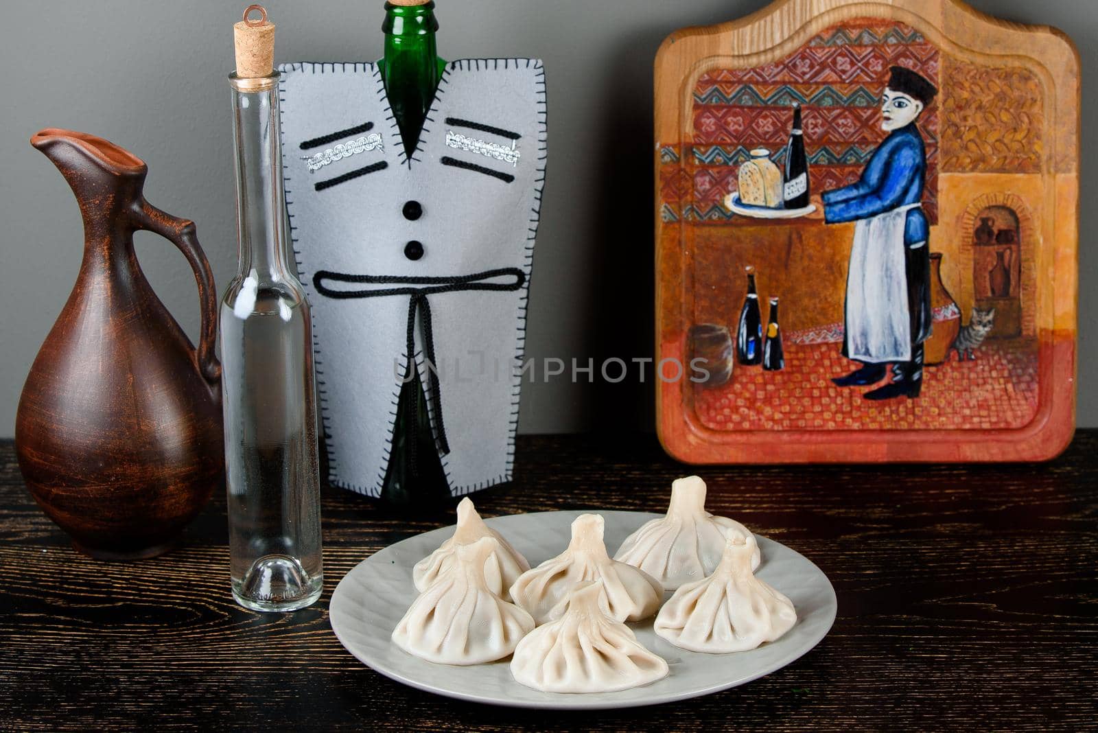 Khinkali on a gray plate against the background of bottles in a Georgian-style decoration, a jug, a board with a thematic pattern on a dark wooden table by Rabizo