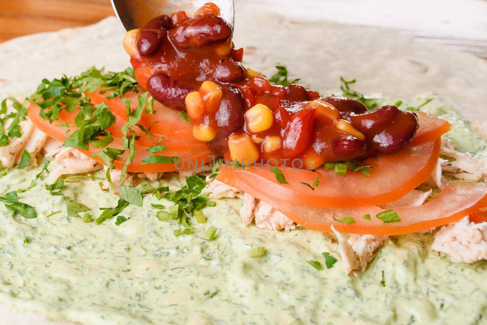 Burrito making process. The cook adds beans and corn in tomato sauce to meat, herbs and tomatoes on pita with sauce. Mexican dish.