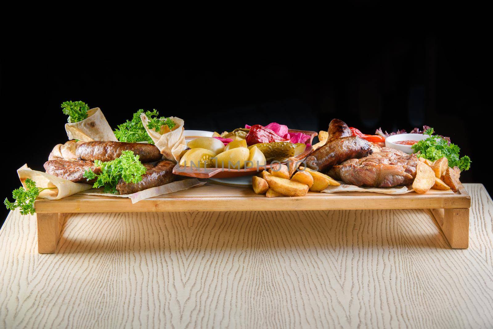 Assortment of fried meat, potatoes, sausages, pickles, tomatoes, peppers, herbs, lavash on a wooden tray on the table.