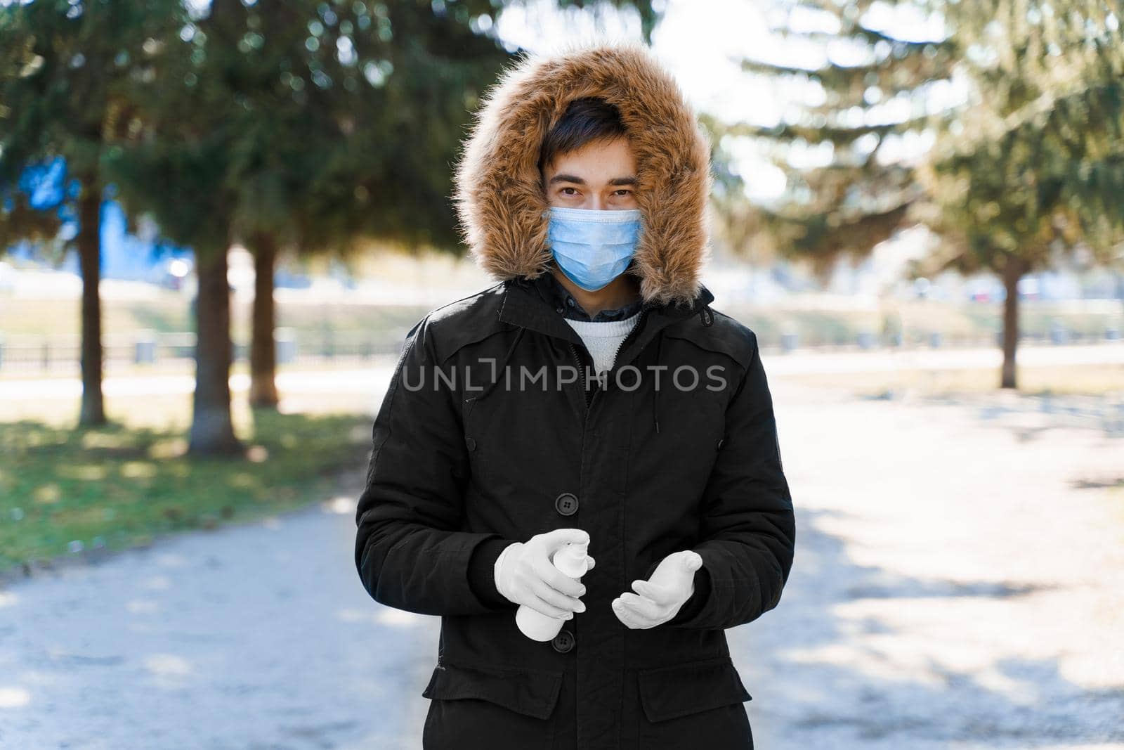 Man sprays antiseptic on his hands which in white medical gloves. Caucasian face in blue sterile medical mask to protect from coronavirus covid 19. by Rabizo
