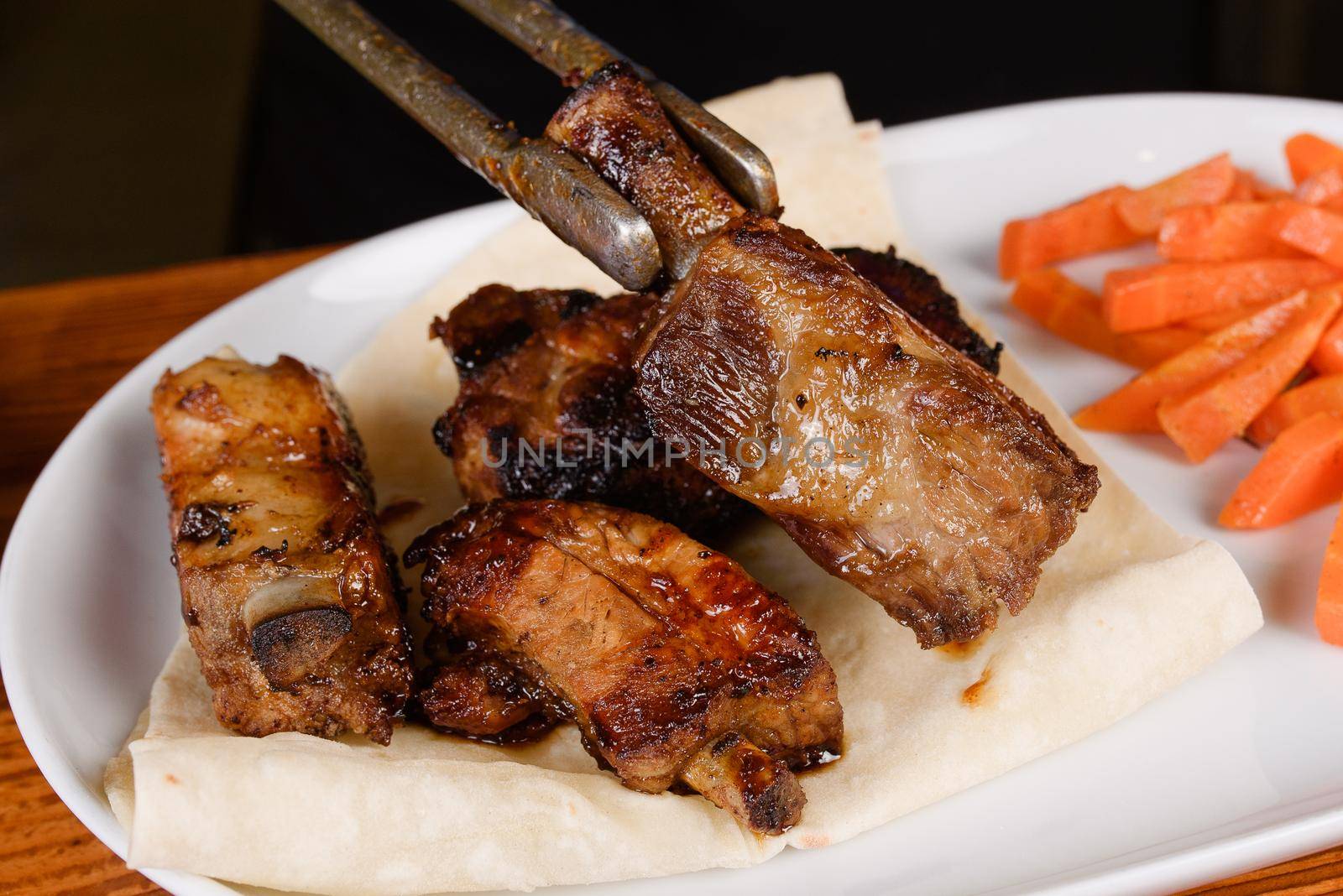 Sliced grilled pork ribs on pita bread with grilled carrots on a white plate on a wooden background by Rabizo