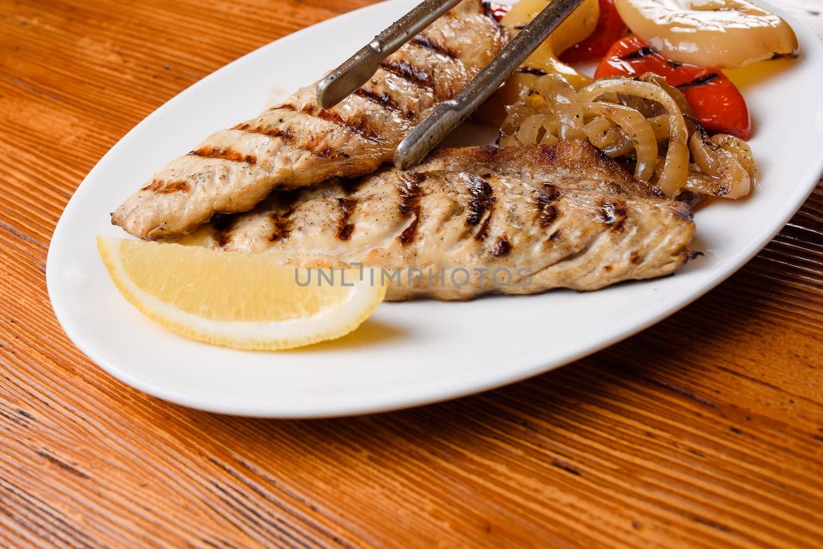 Chicken fillet and grilled vegetables with lemon on a white plate on a wooden table. Serving the dish with metal tongs.