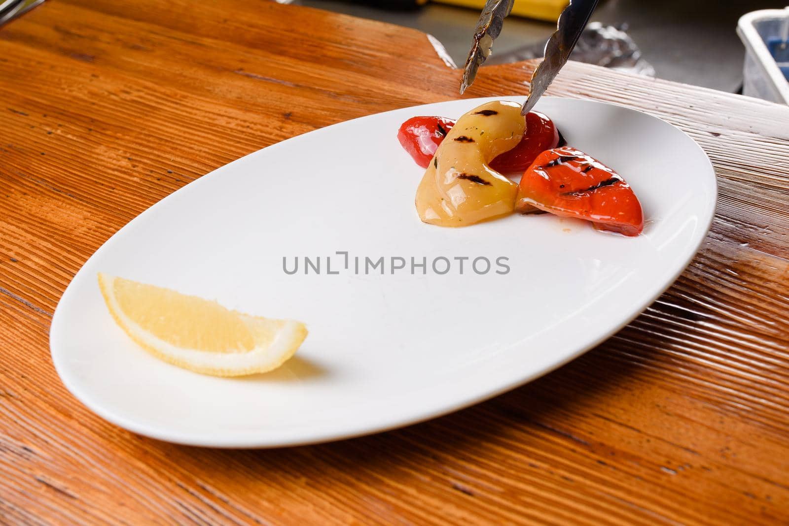 Grilled pepper and with lemon on a white plate on a wooden table. Serving the dish with metal tongs.