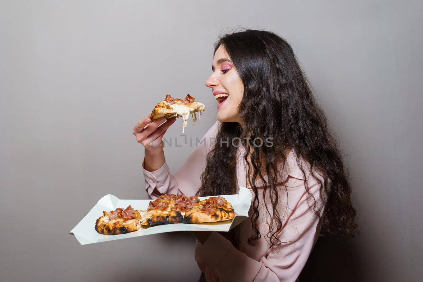 Girl courier eating pinsa pizza romana gourmet italian cuisine on grey background. Holding scrocchiarella traditional dish. Food delivery from pizzeria. Pinsa with meat, arugula, olives, cheese.