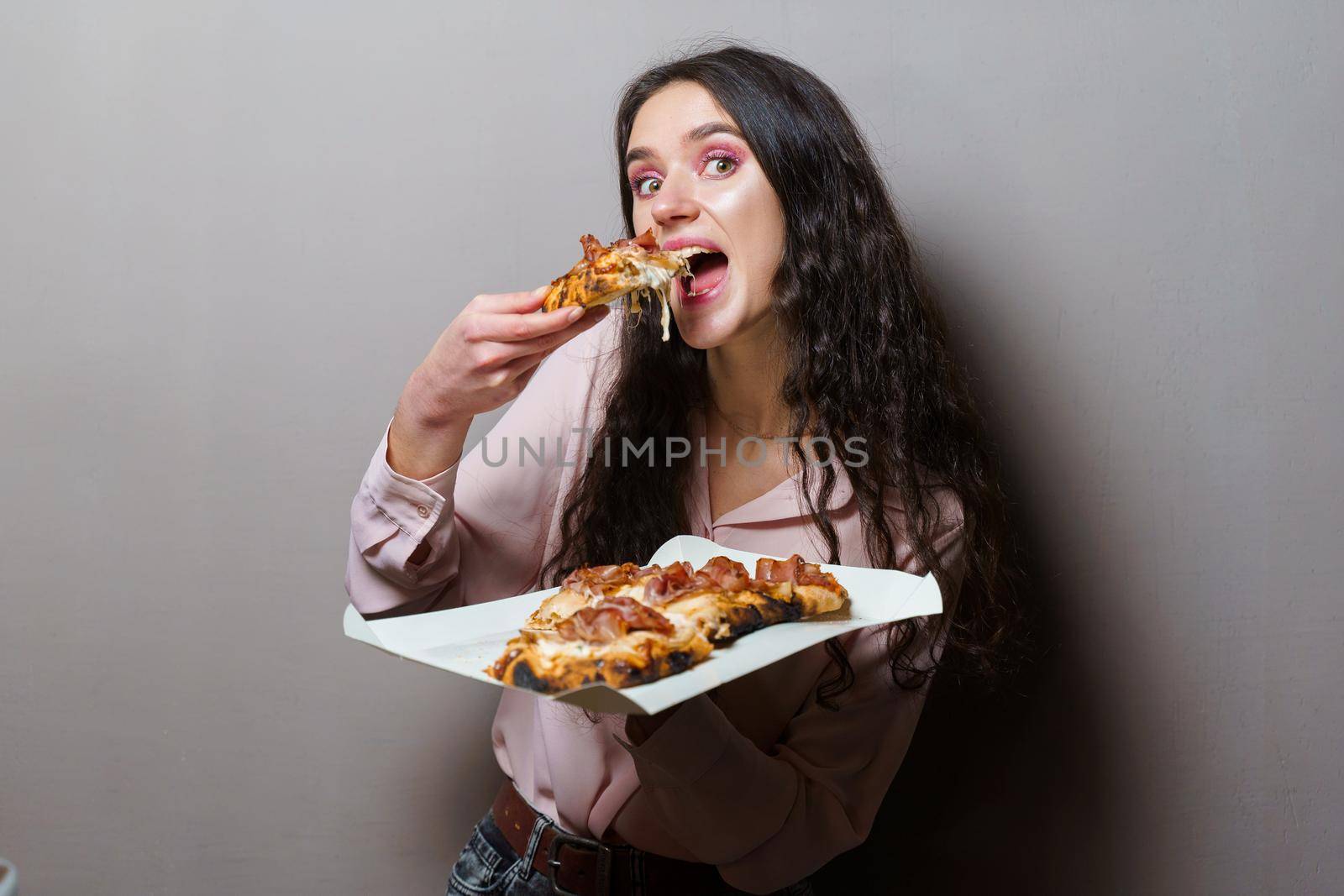 Girl courier eating pinsa pizza romana gourmet italian cuisine on grey background. Holding scrocchiarella traditional dish. Food delivery from pizzeria. Pinsa with meat, arugula, olives, cheese.