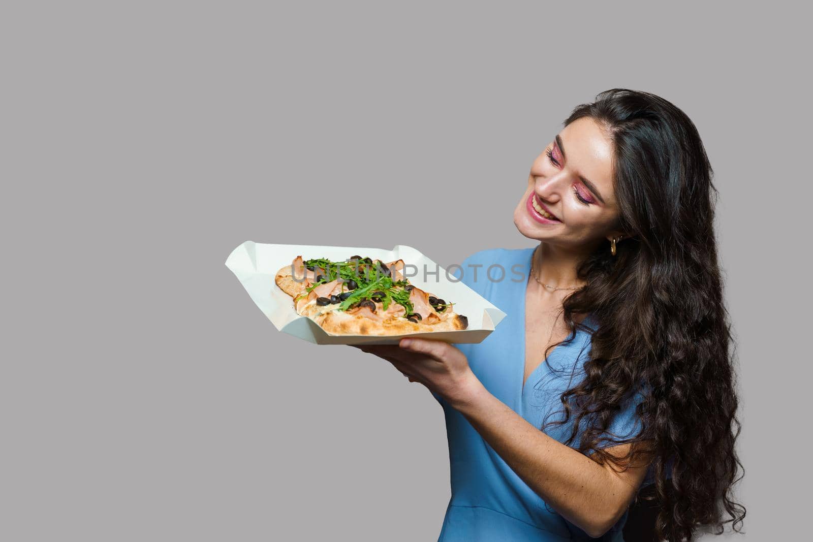Girl courier with pinsa romana gourmet italian cuisine on grey background. Holding scrocchiarella traditional dish. Food delivery from pizzeria. Pinsa with meat, arugula, olives, cheese