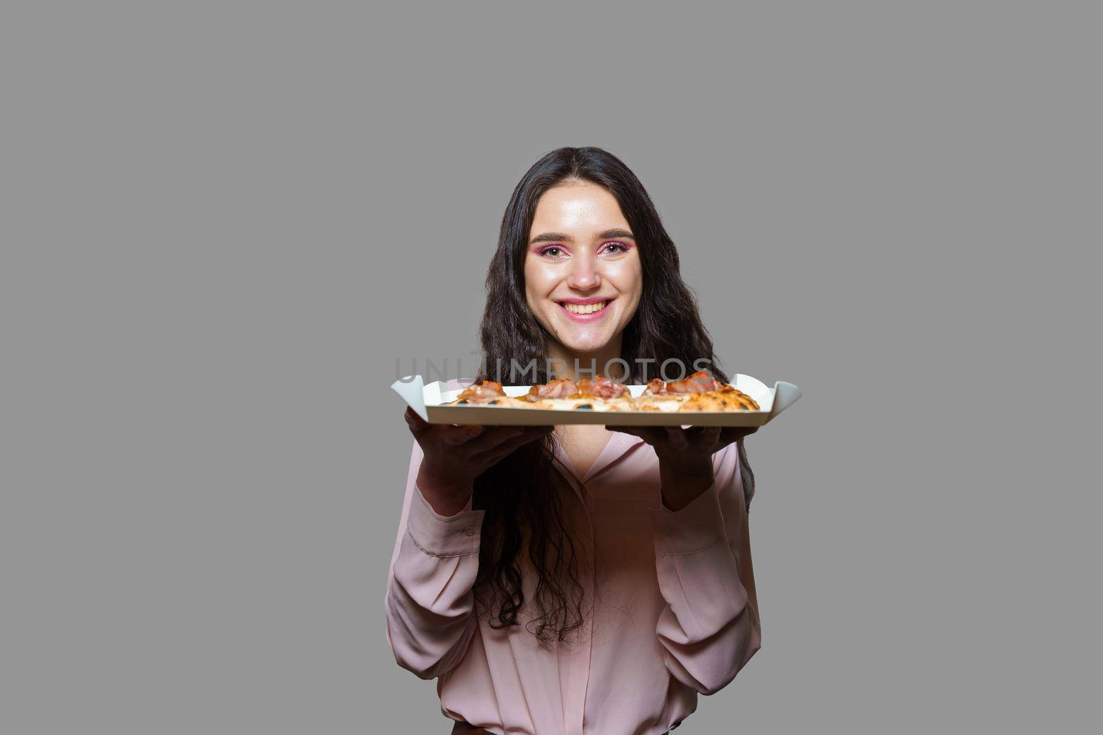 Girl courier with pinsa romana gourmet italian cuisine on grey background. Holding scrocchiarella traditional dish. Food delivery from pizzeria. Pinsa with meat, arugula, olives, cheese.