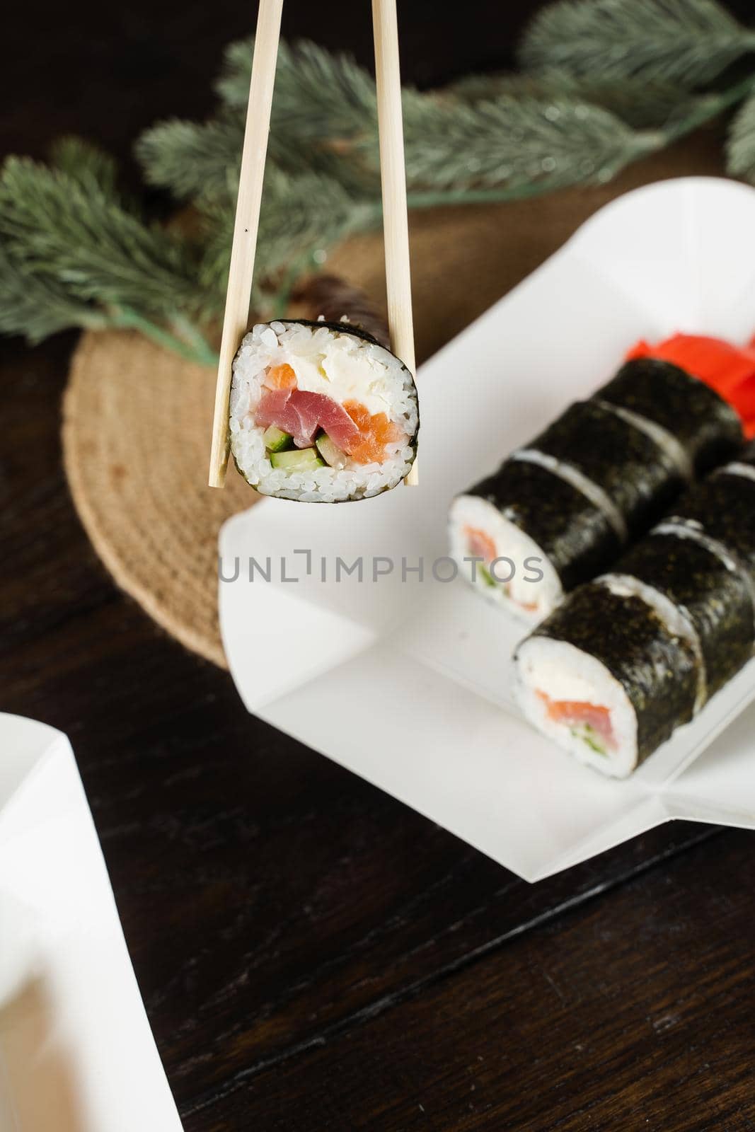 Holding sushi roll using chopsticks on the food delivery container. Christmas decoration. Food delivery at new year eve. Roll with salmon, tuna, cucumber and soft cheese