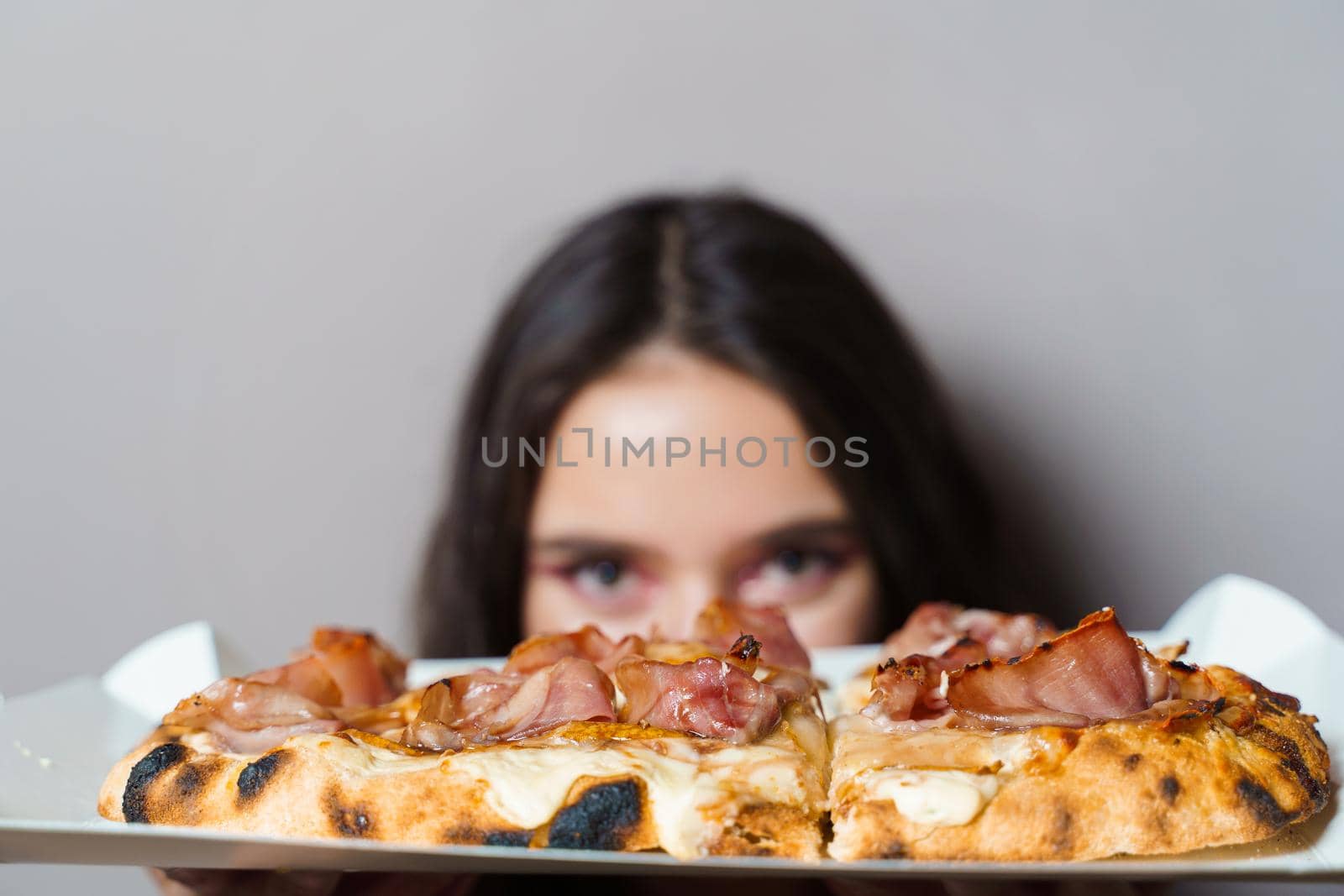 Girl courier with pinsa romana gourmet italian cuisine on grey background. Holding scrocchiarella traditional dish. Food delivery from pizzeria. Pinsa with meat, arugula, olives, cheese.