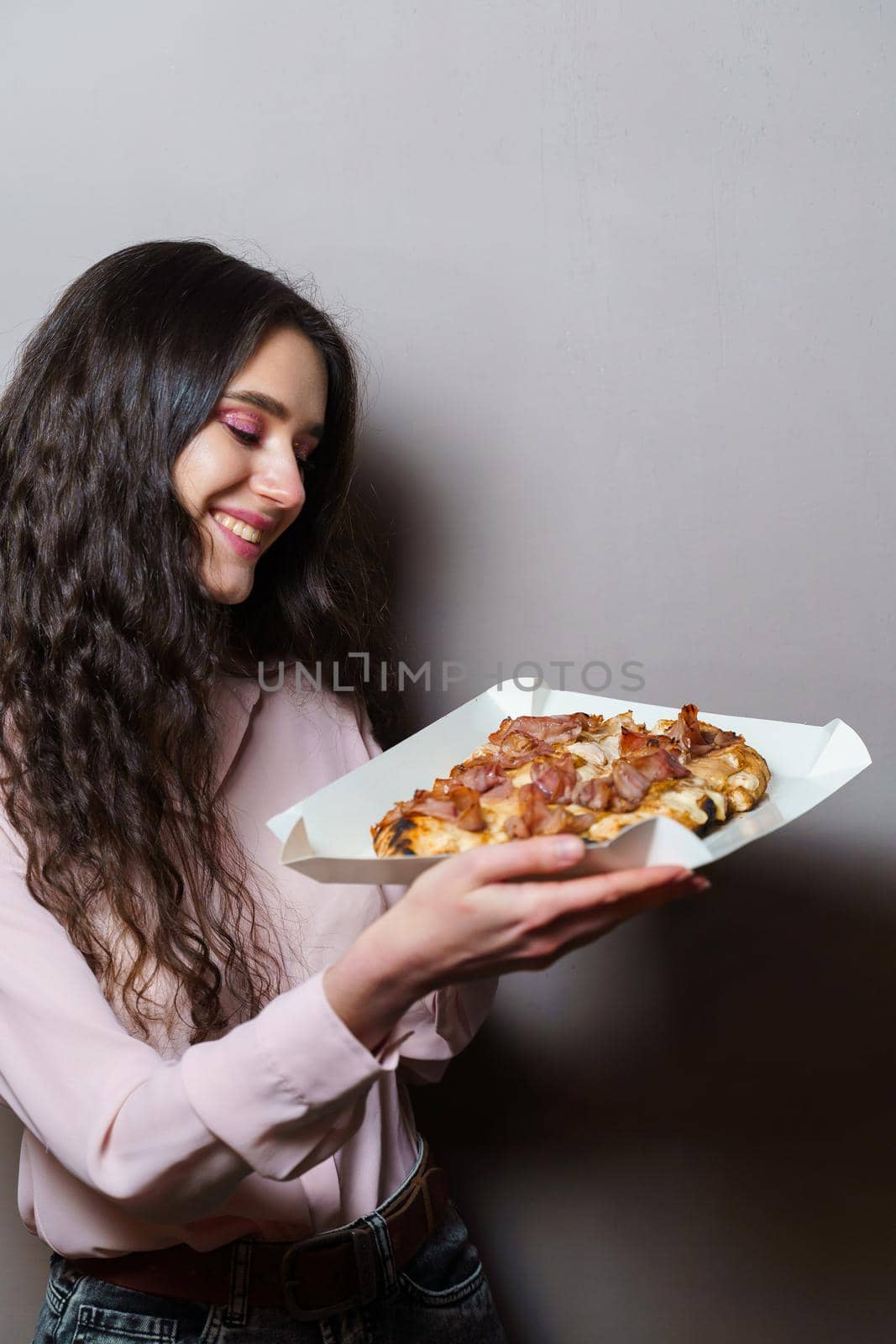 Girl courier with pinsa romana gourmet italian cuisine on grey background. Holding scrocchiarella traditional dish. Food delivery from pizzeria. Pinsa with meat, arugula, olives, cheese.