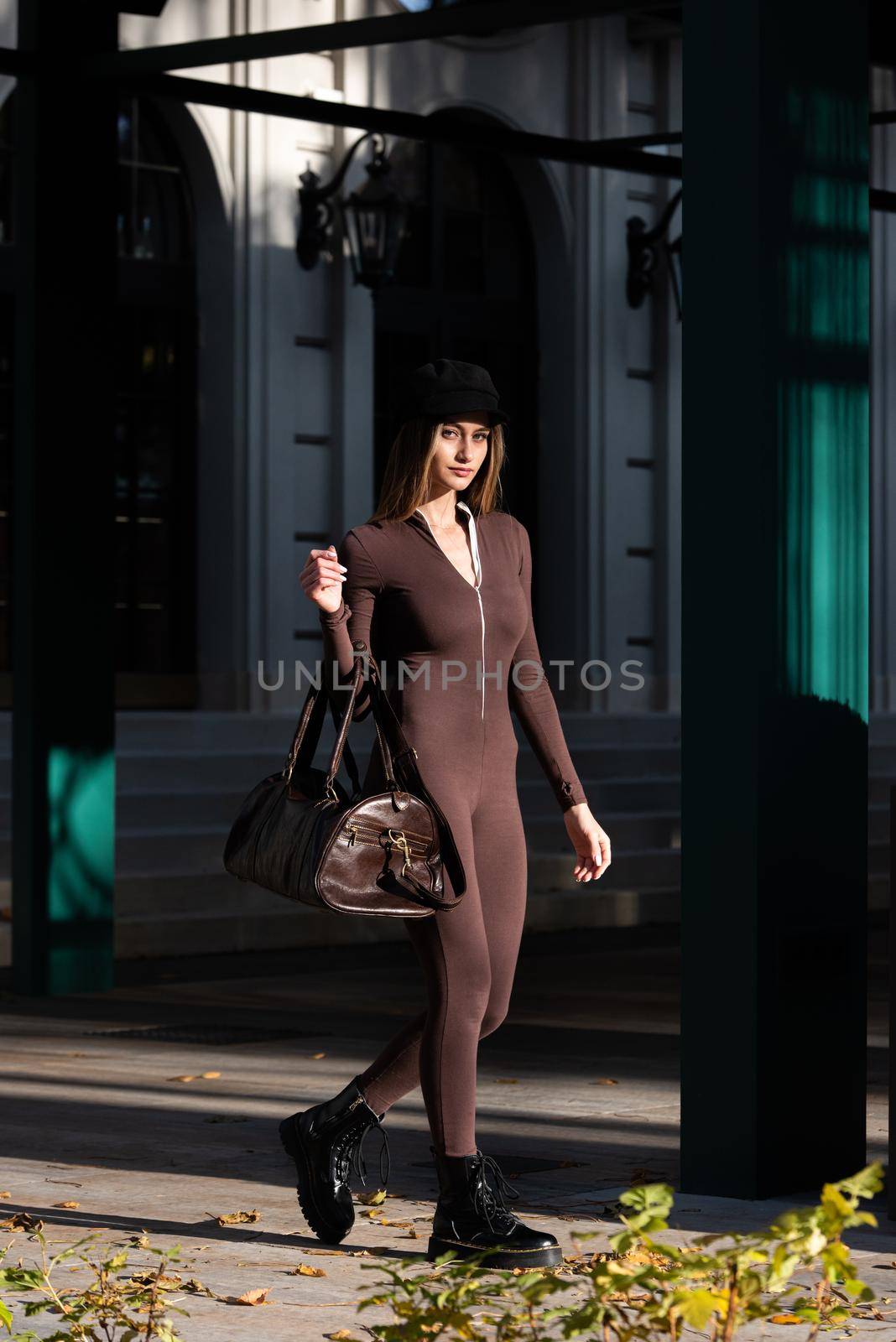 Fashionable young brunette woman with long legs. wearing Solid Long Sleeve Bodycon One Piece Jumpsuits posing with a leather brown travel bag on city street in old town