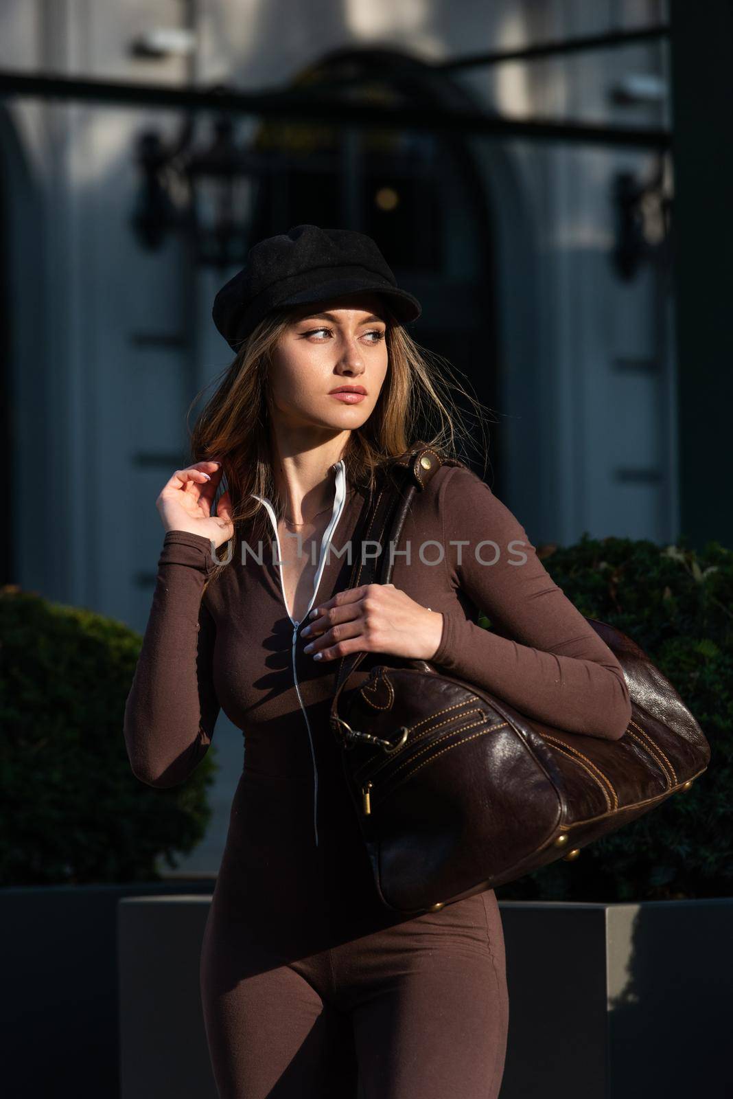 Fashionable young brunette woman with long legs. wearing Solid Long Sleeve Bodycon One Piece Jumpsuits posing with a leather brown travel bag on city street in old town.