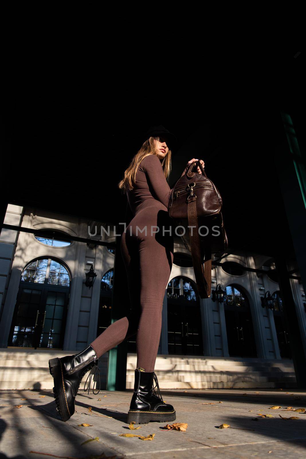 Fashionable young brunette woman with long legs. wearing Solid Long Sleeve Bodycon One Piece Jumpsuits posing with a leather brown travel bag on city street in old town