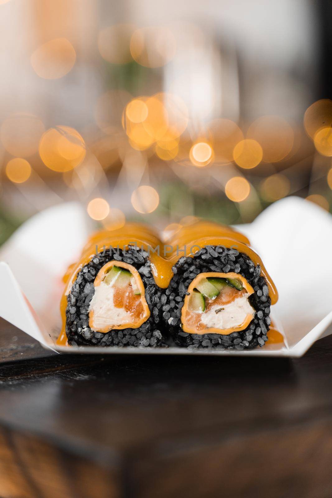 Sushi black roll on the new year lights background. Christmas lights decoration. Food delivery at new year eve. Roll with black rice, salmon, cucumber and cheese