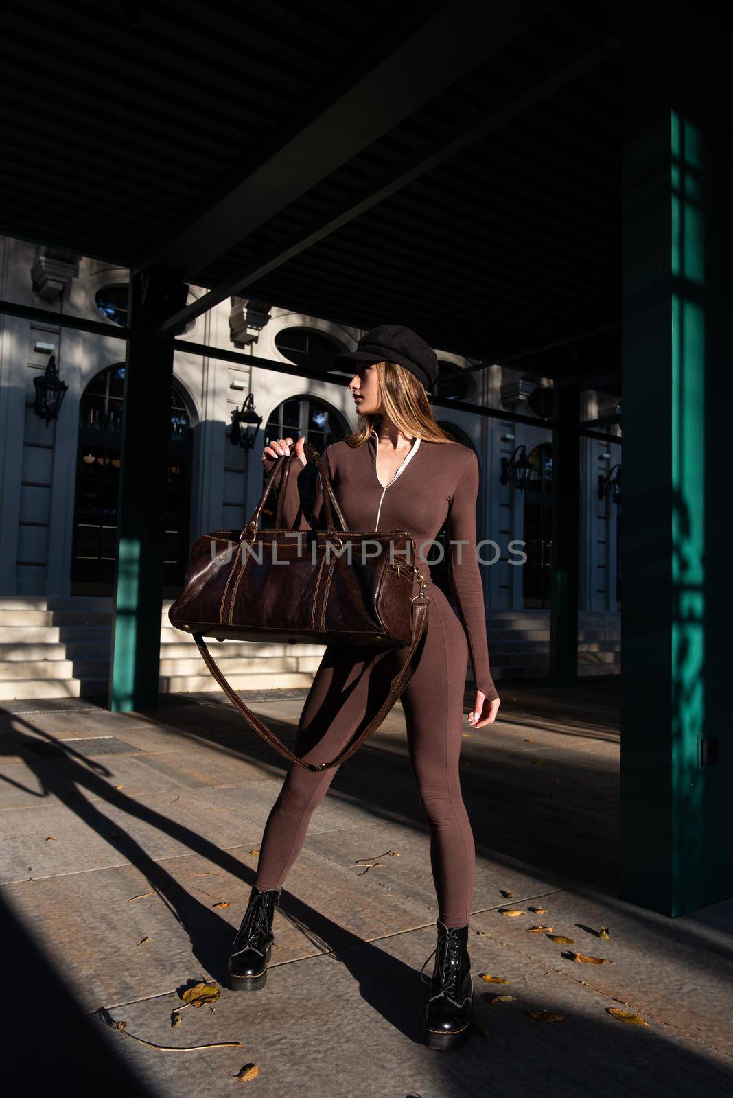 Fashionable young brunette woman with long legs. wearing Solid Long Sleeve Bodycon One Piece Jumpsuits posing with a leather brown travel bag on city street in old town