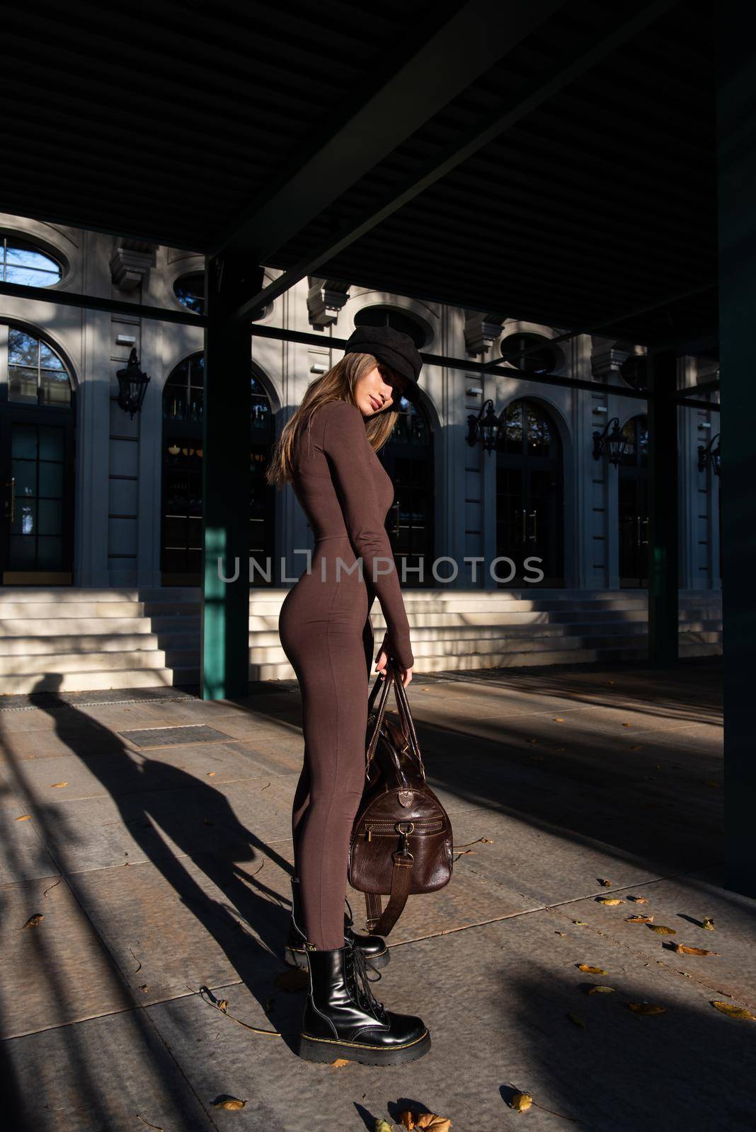 Fashionable young brunette woman with long legs. wearing Solid Long Sleeve Bodycon One Piece Jumpsuits posing with a leather brown travel bag on city street in old town